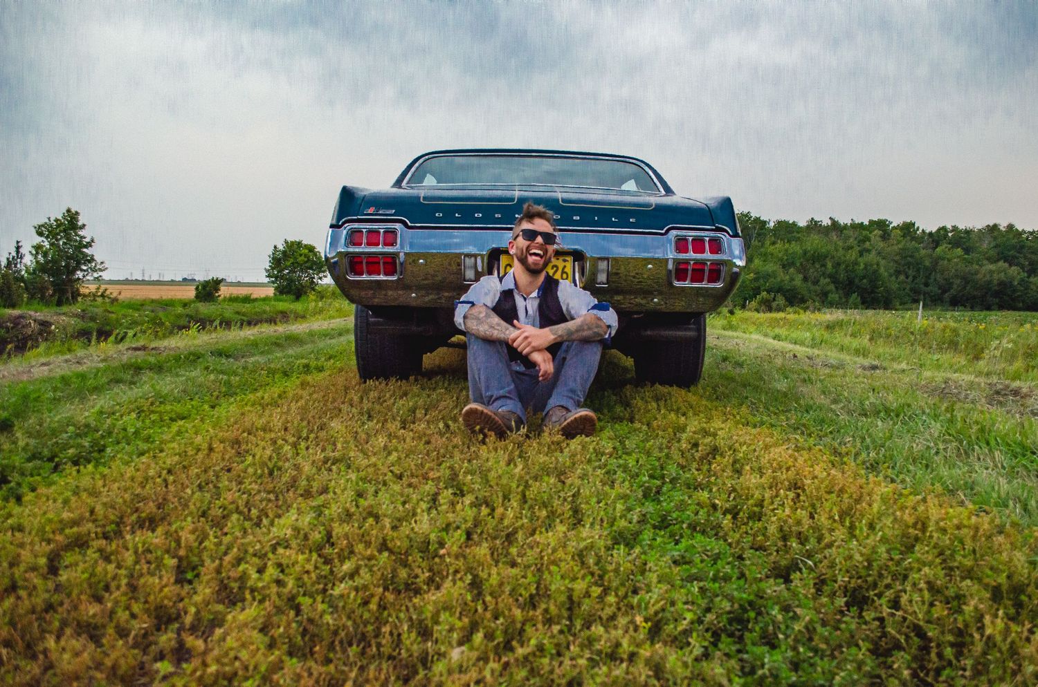 Man in sunglasses sitting in grass behind a vintage blue classic car in a rural field on a cloudy day.
