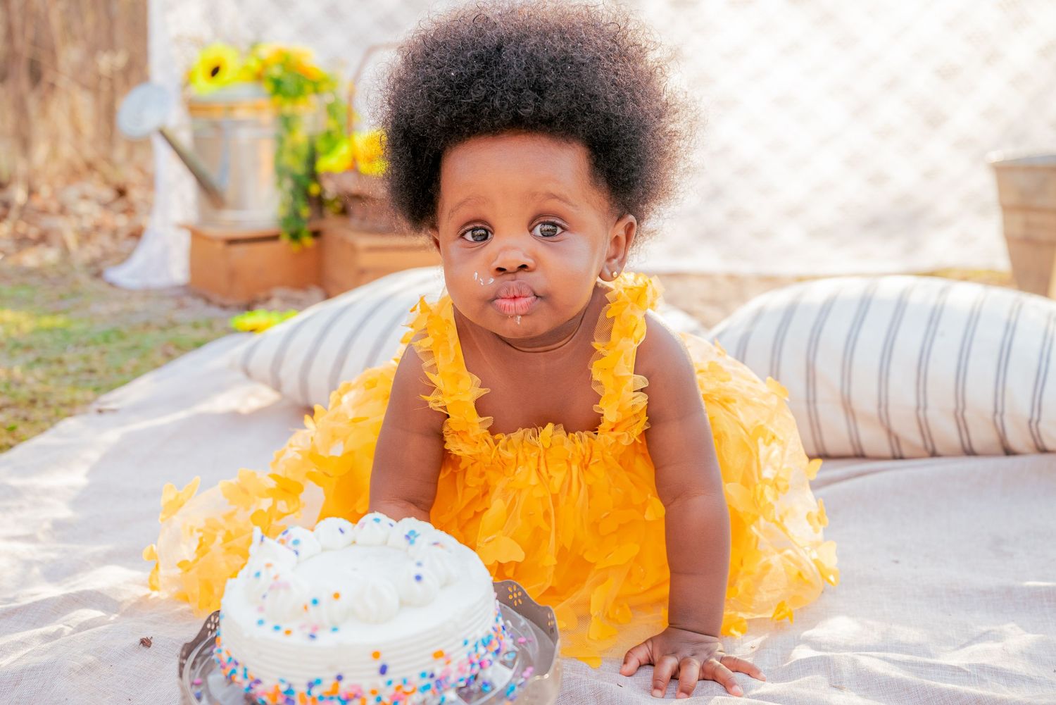 Adorable baby girl in yellow ruffle dress sitting near white birthday cake with colorful sprinkles outdoors.