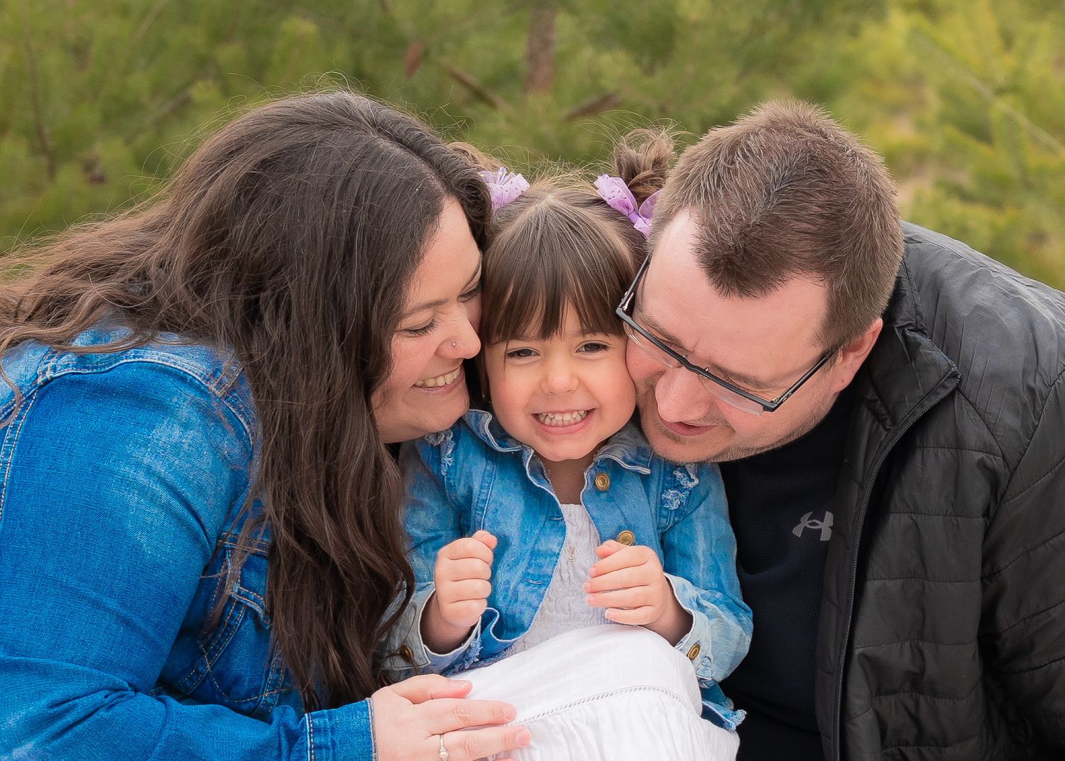 Parents lovingly kissing their young daughter on the cheeks outdoors in a pine forest.