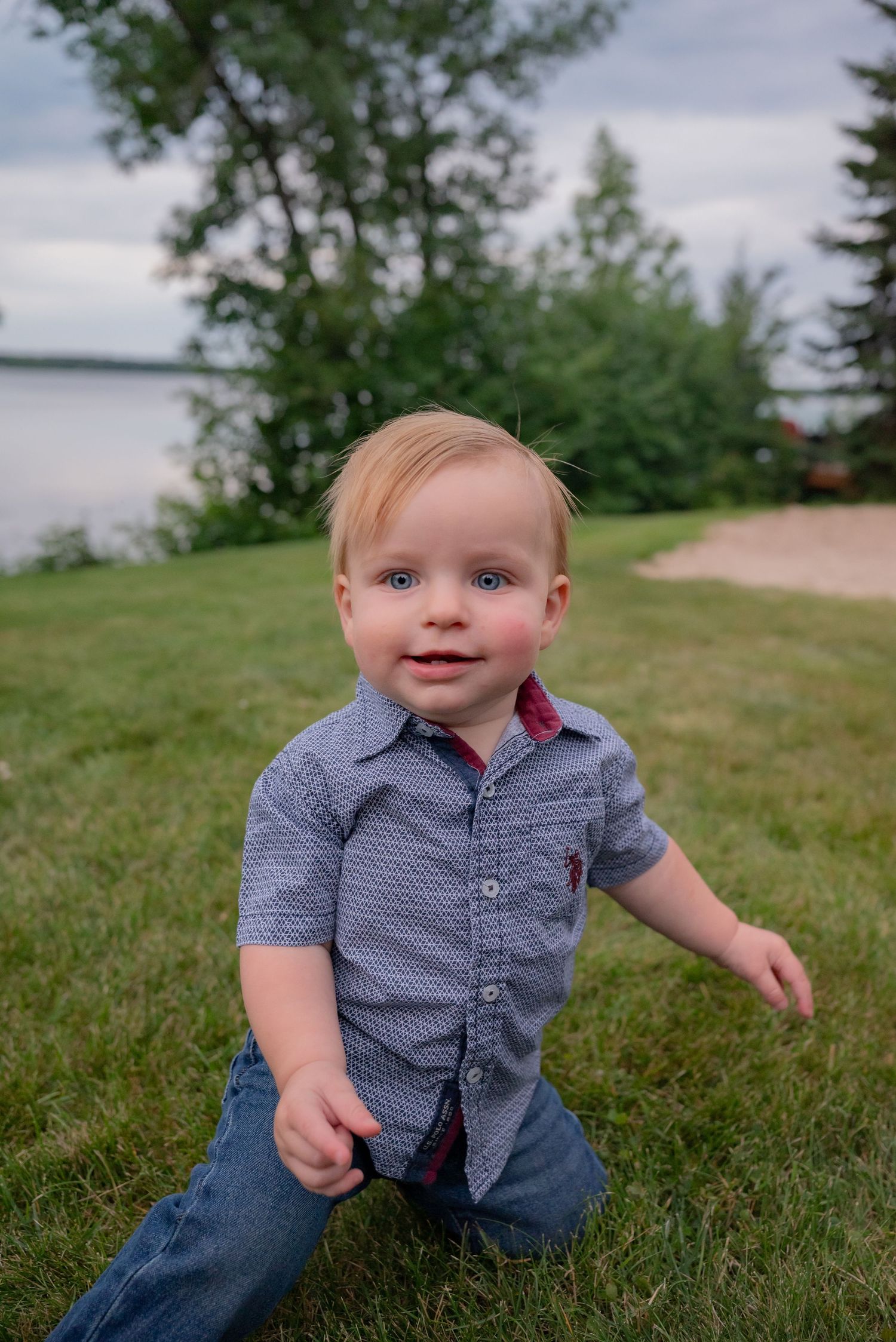 Smiling baby boy with blue eyes wearing a blue button-up shirt and jeans sitting on green grass near a lake.