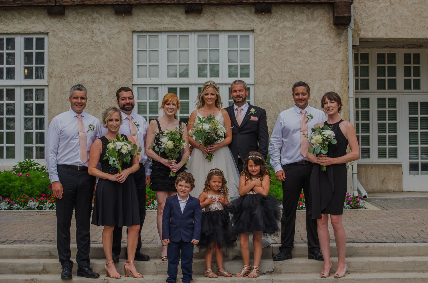 Wedding party poses on stone steps, bride and groom centered with bridesmaids, groomsmen, and children in formal attire.