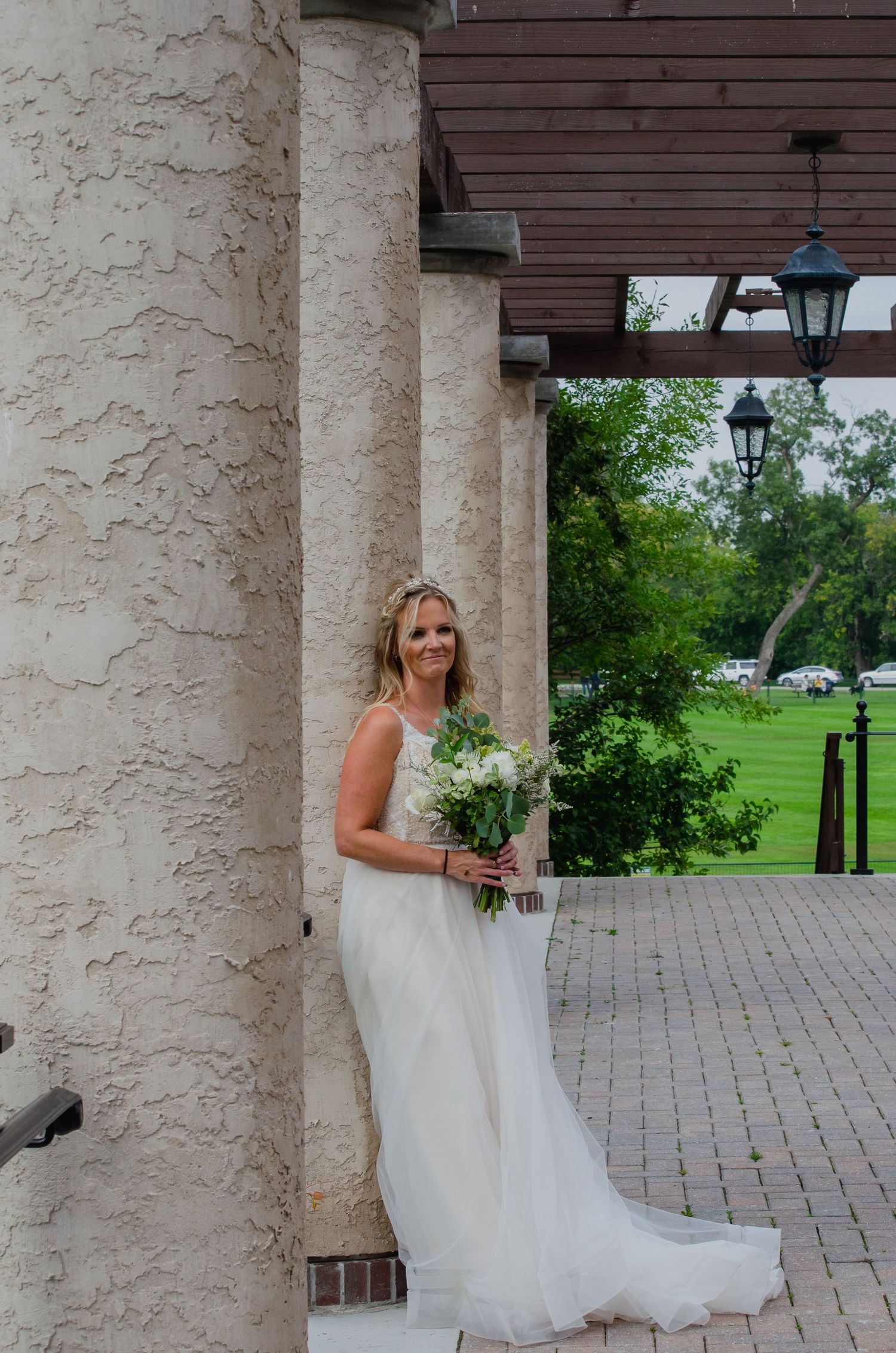 Bride in white gown holding bouquet of white flowers, leaning against stone column at elegant outdoor venue.