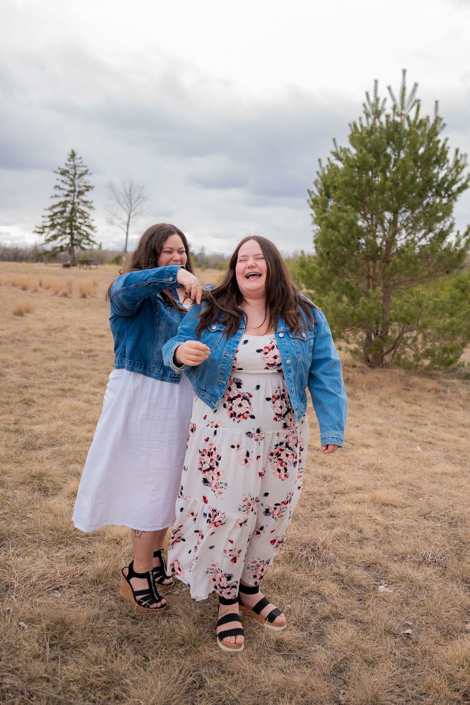 Two women in floral dresses and denim jackets laughing outdoors in a field with trees and cloudy sky.