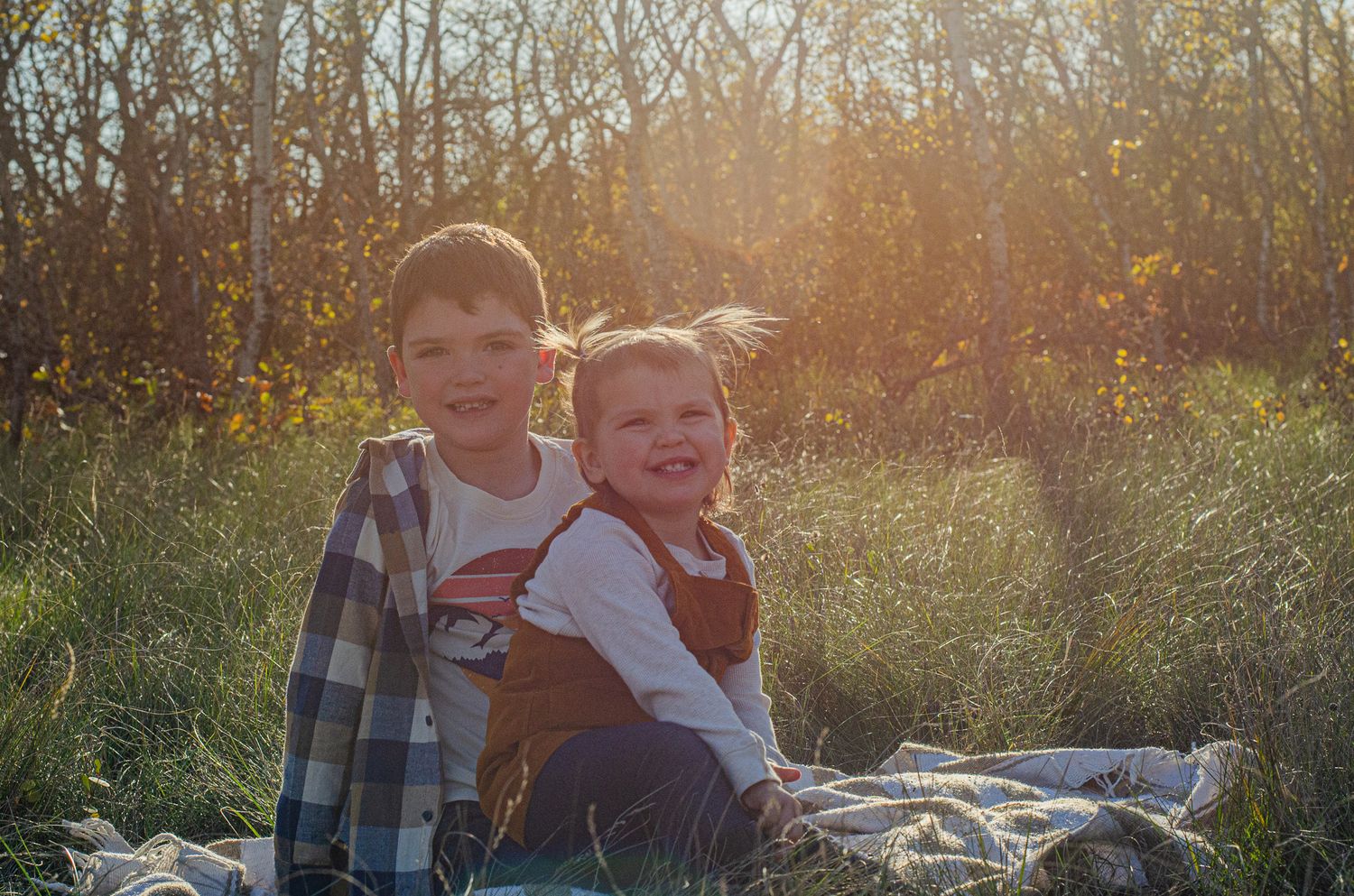 Two happy children sitting on a blanket in a sunny field, smiling together during a golden hour outdoor photo session.