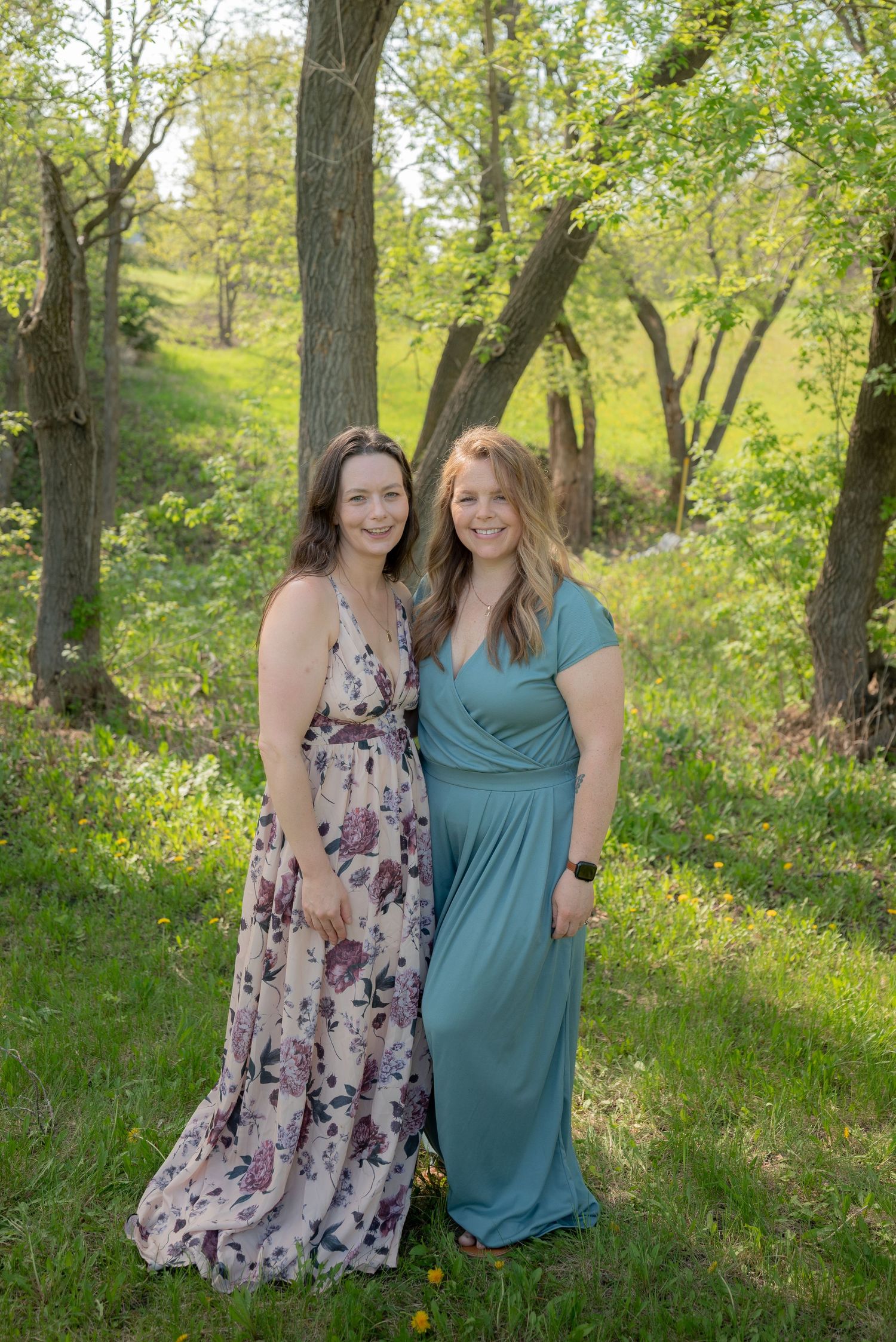 Two women in elegant maxi dresses pose outdoors in a lush green wooded area during spring.