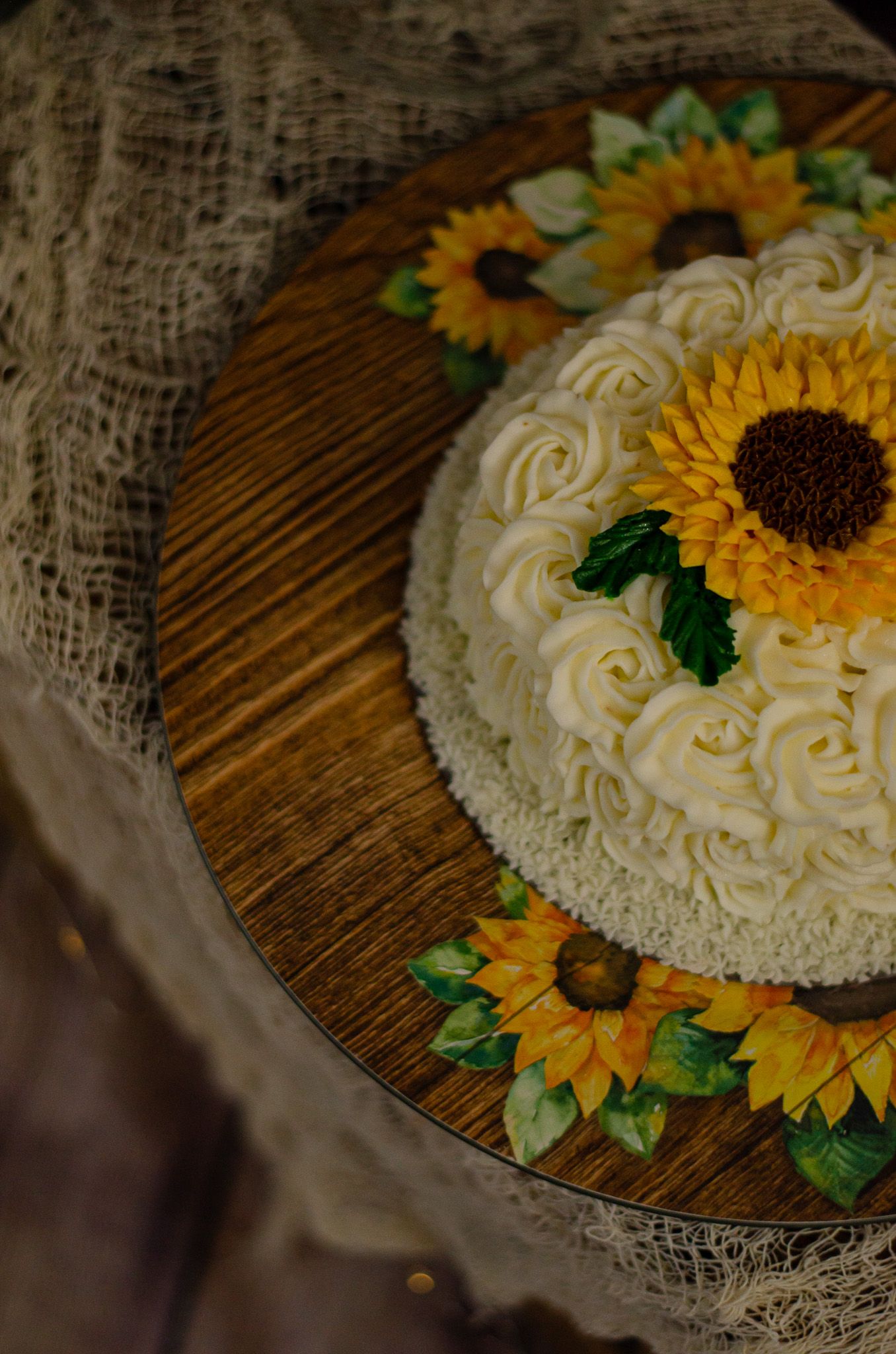 White rosette cake topped with a sunflower on a wooden stand, surrounded by sunflower decorations and burlap.