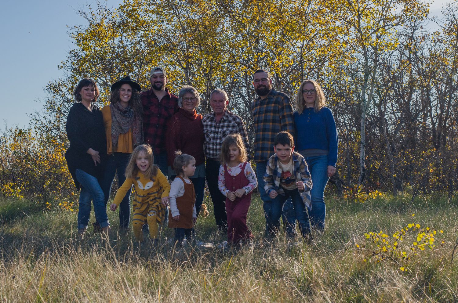 Family group photo outdoors in autumn field with golden trees, adults and children posing together in fall clothing.