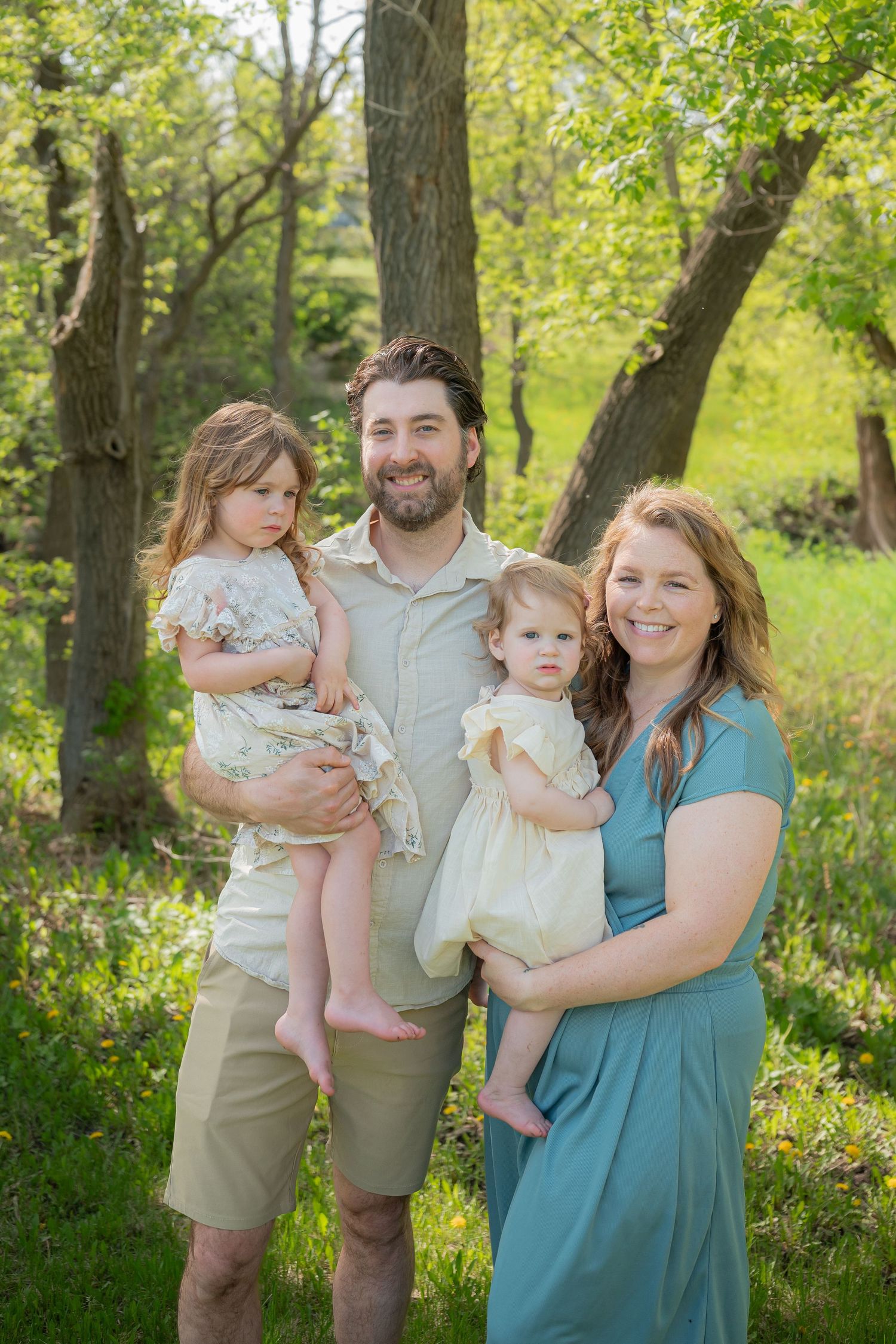 Happy family of four posing outdoors in lush green park, parents holding two young daughters in floral dresses.