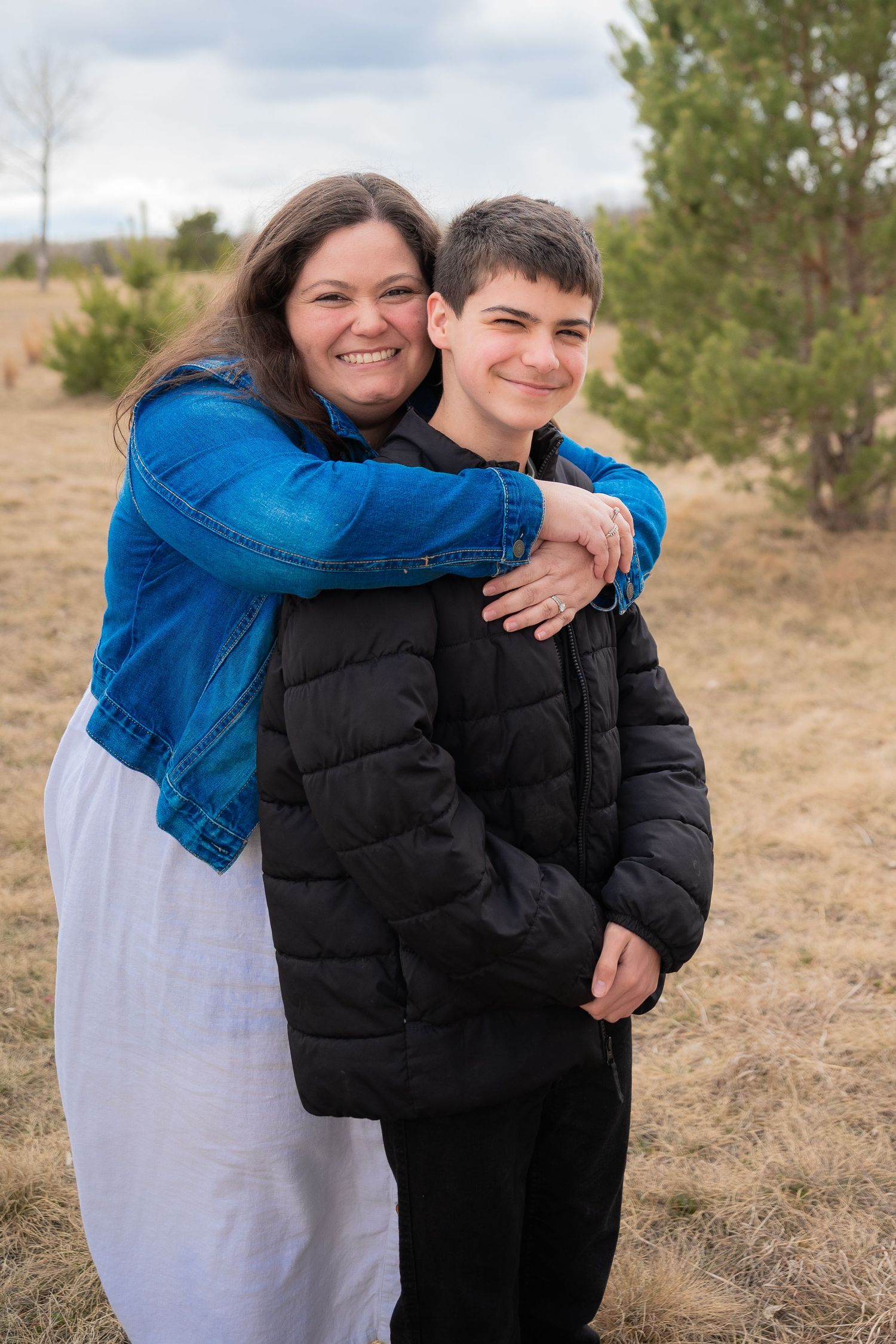 A smiling woman in a blue denim jacket hugs a teenage boy in a black puffer jacket outdoors.