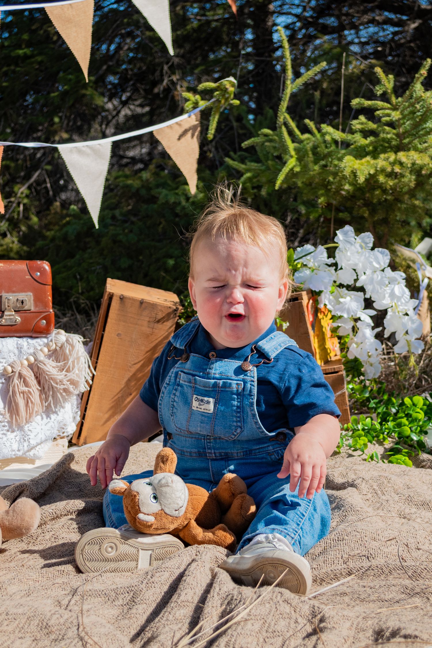 A one-year-old baby in denim overalls crying outdoors at a birthday party setup with bunting and a teddy bear.