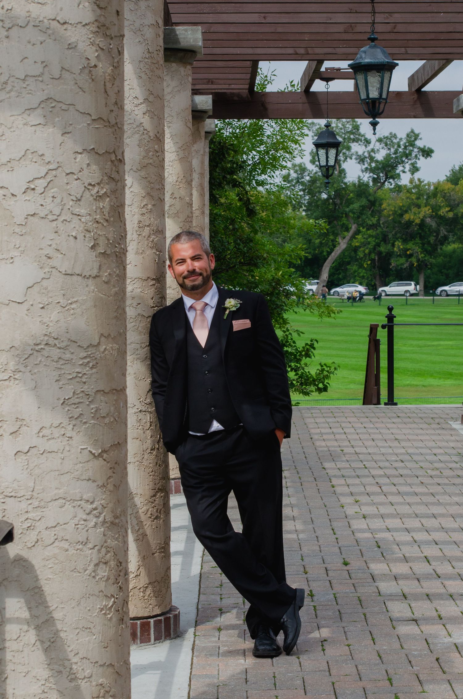 Groom in black suit with pink tie leaning against stone column at outdoor wedding venue with green lawn.