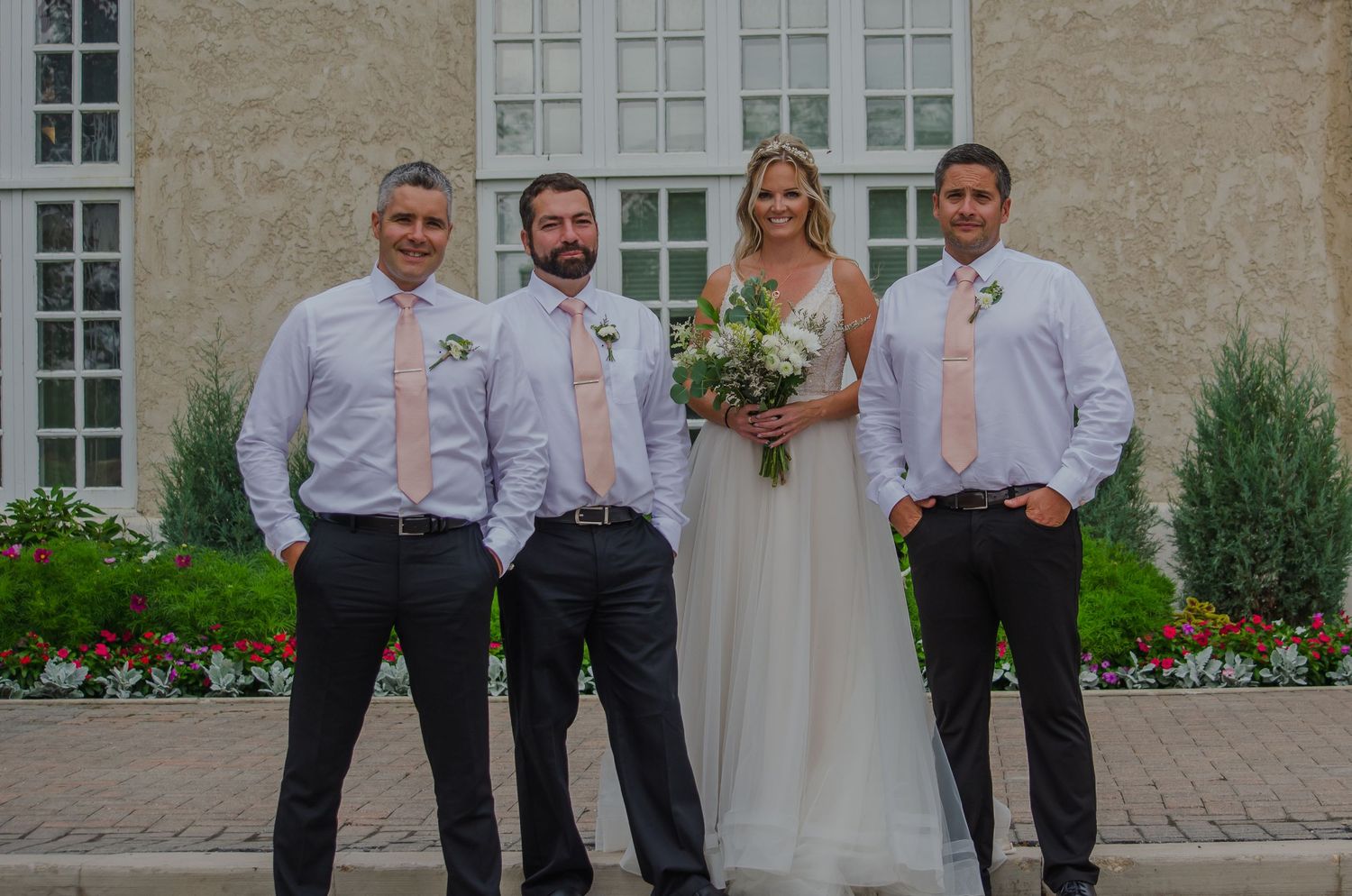 Bride in white gown holding bouquet poses with three groomsmen in pink ties outside elegant stone building.