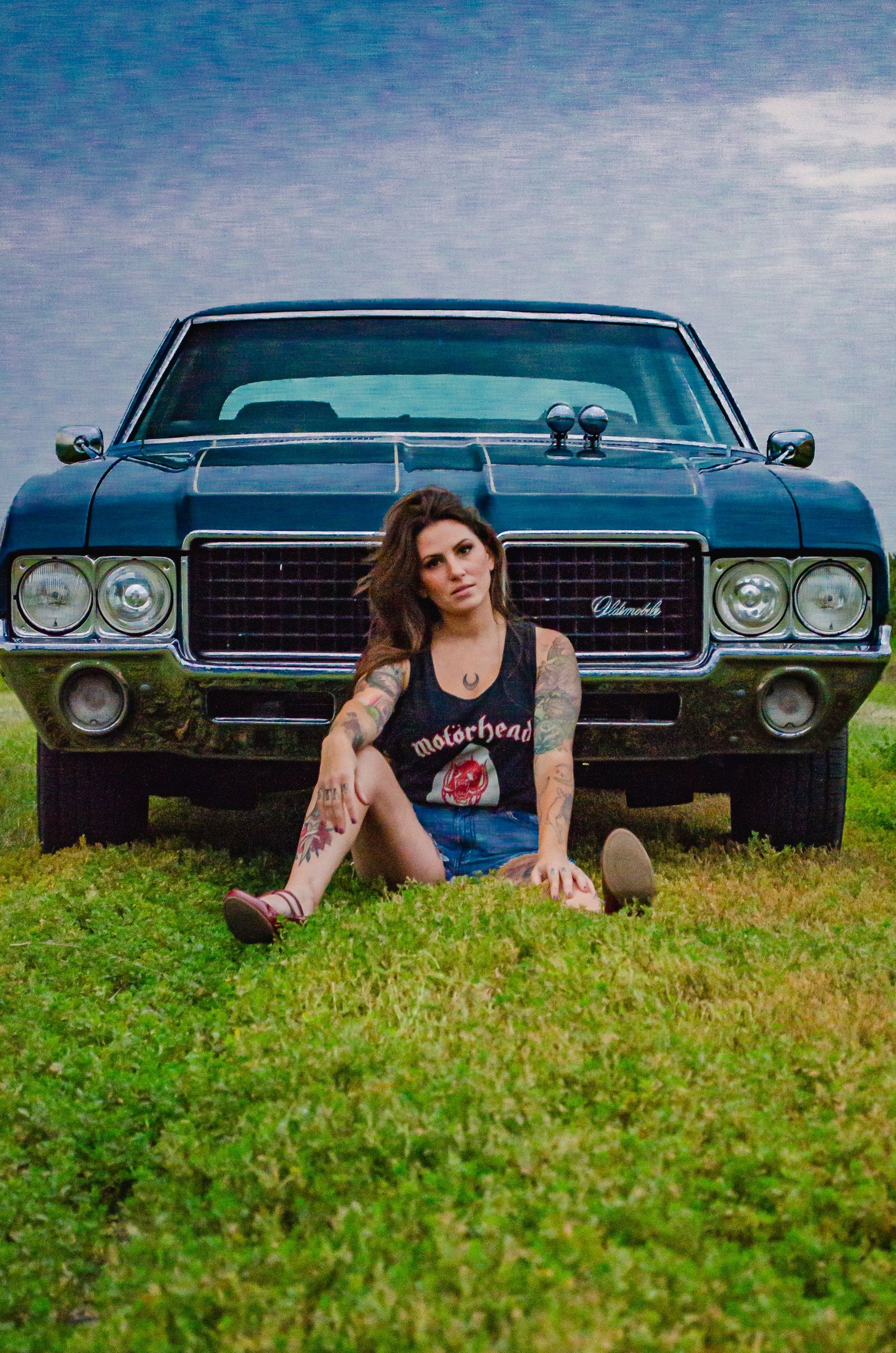 Tattooed woman in Motorhead tank top sitting on grass in front of a classic blue Oldsmobile muscle car.