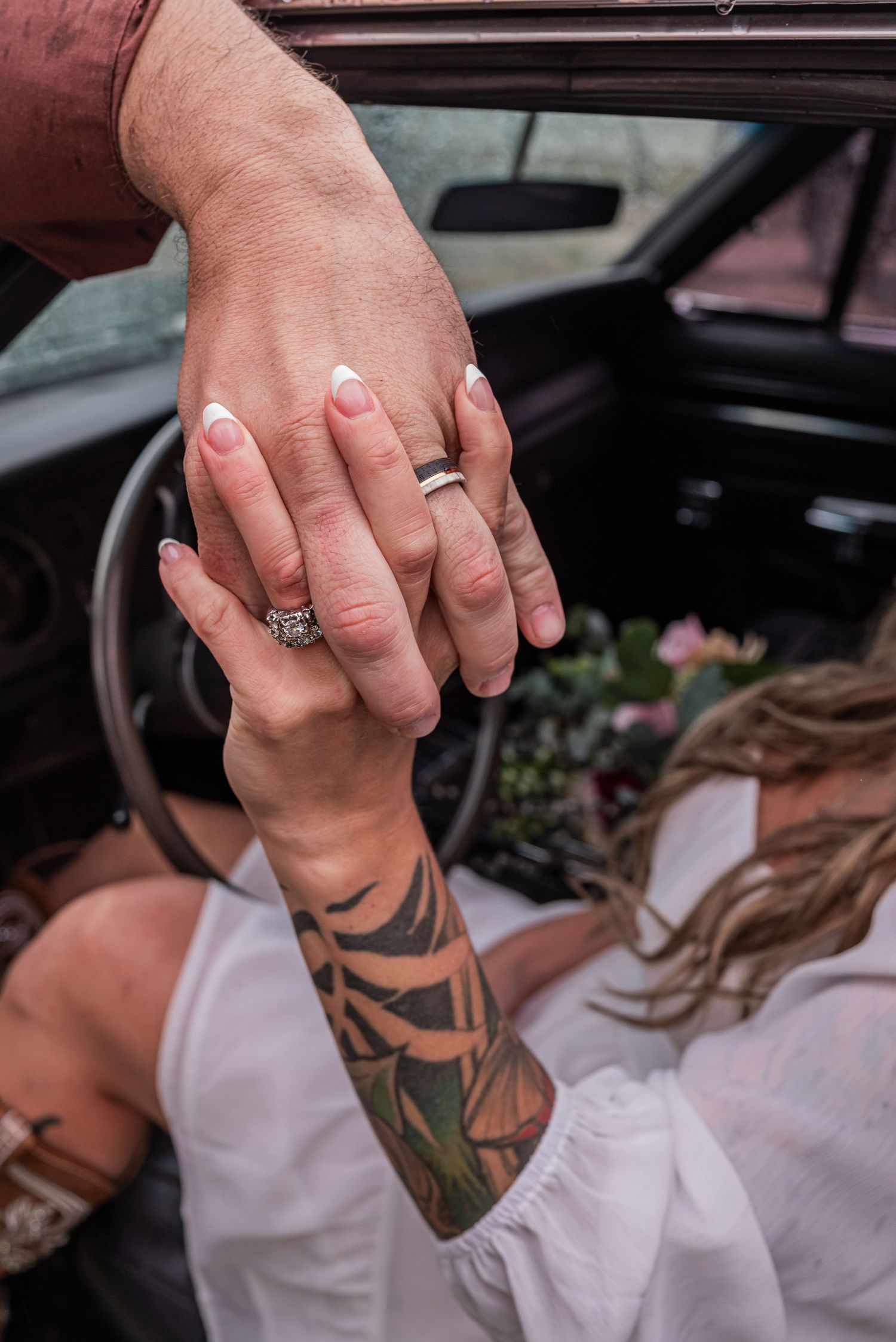 Newlywed couple displaying wedding rings, bride with floral tattoo sleeve in white dress holding bouquet inside vintage car.