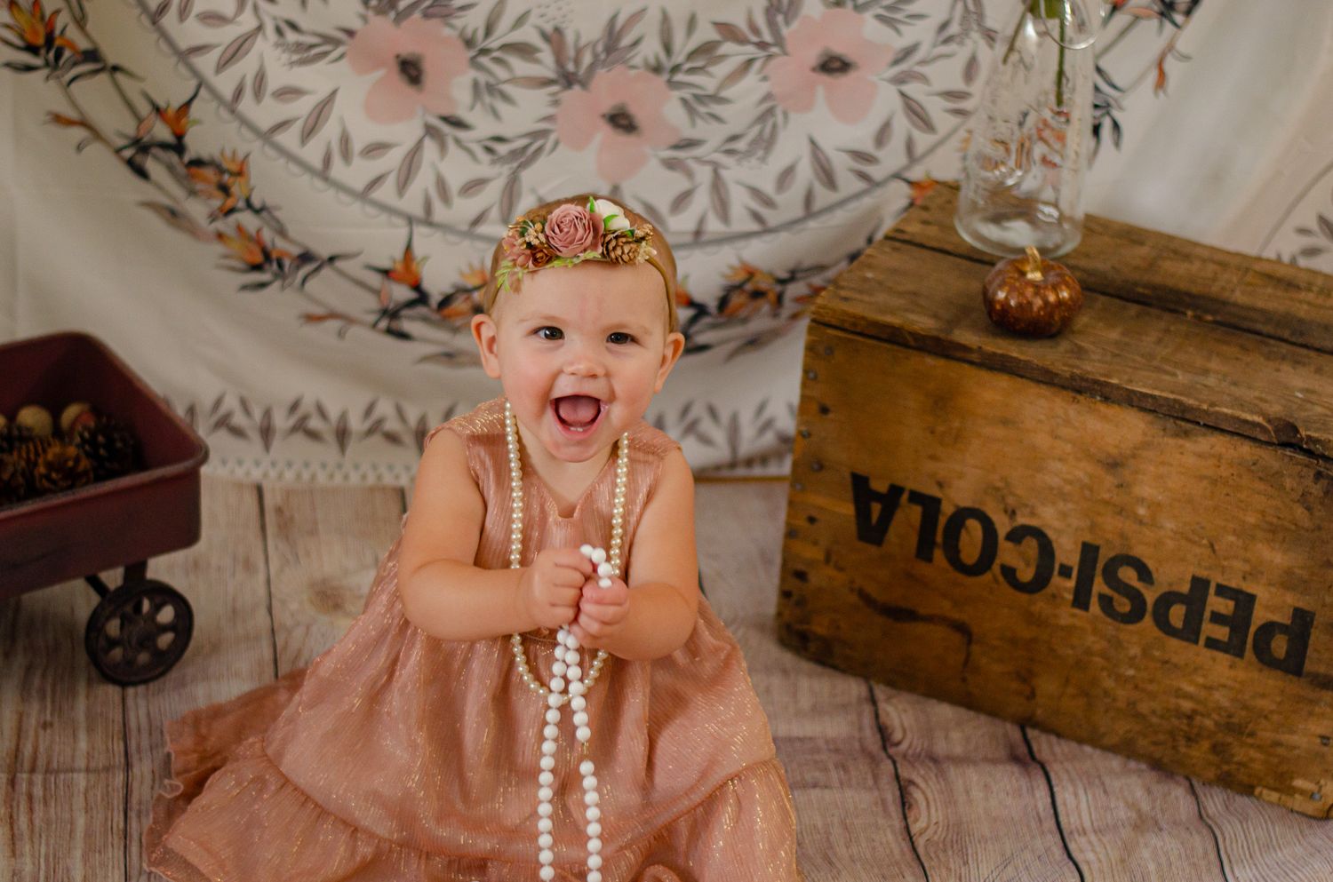 Laughing baby girl in pink dress and pearl necklace with floral headband sits before a Pepsi-Cola crate.
