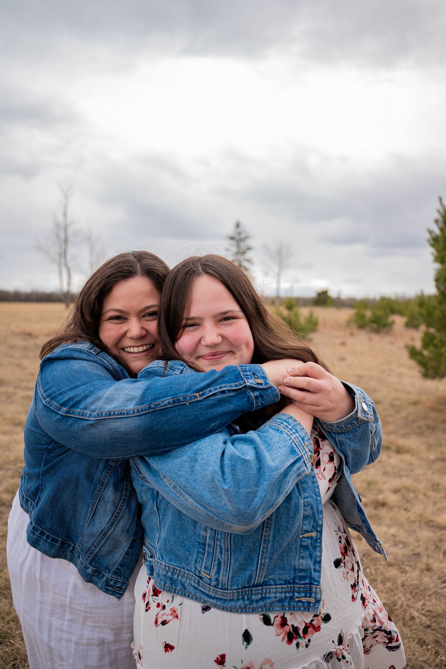 Two young women in denim jackets share a warm hug outdoors in a field under a cloudy sky.
