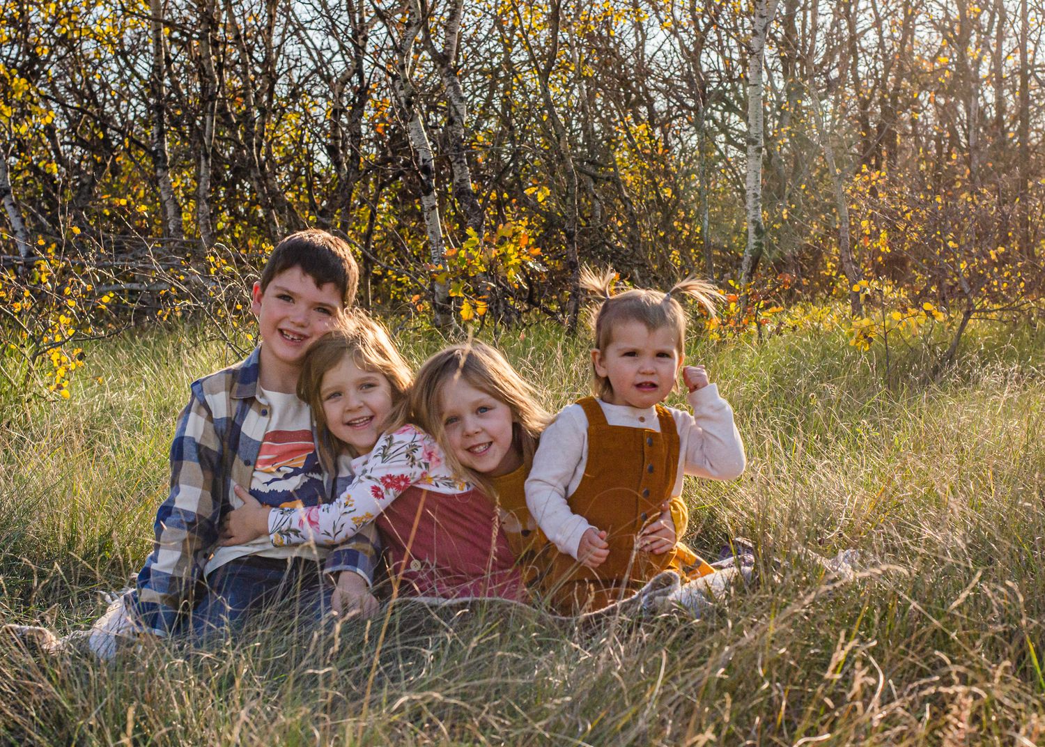 Four children smile together in a sunny autumn field surrounded by golden grass and yellow wildflowers.