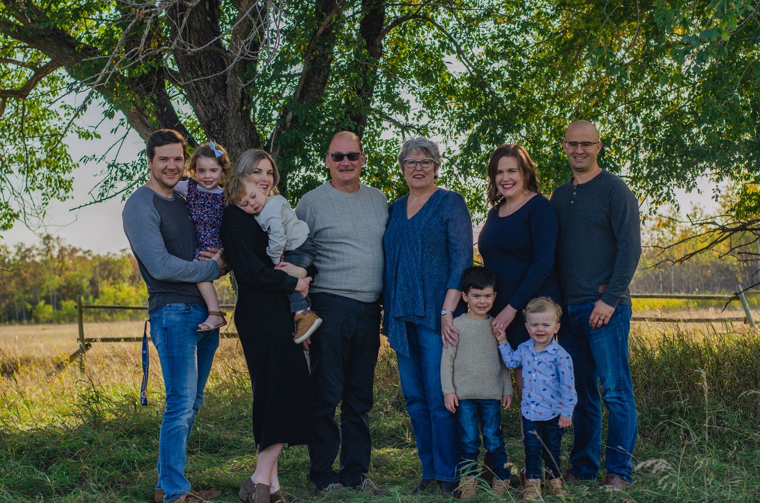Multi-generational family of eight smiling together outdoors under a large tree in a scenic rural field at golden hour.