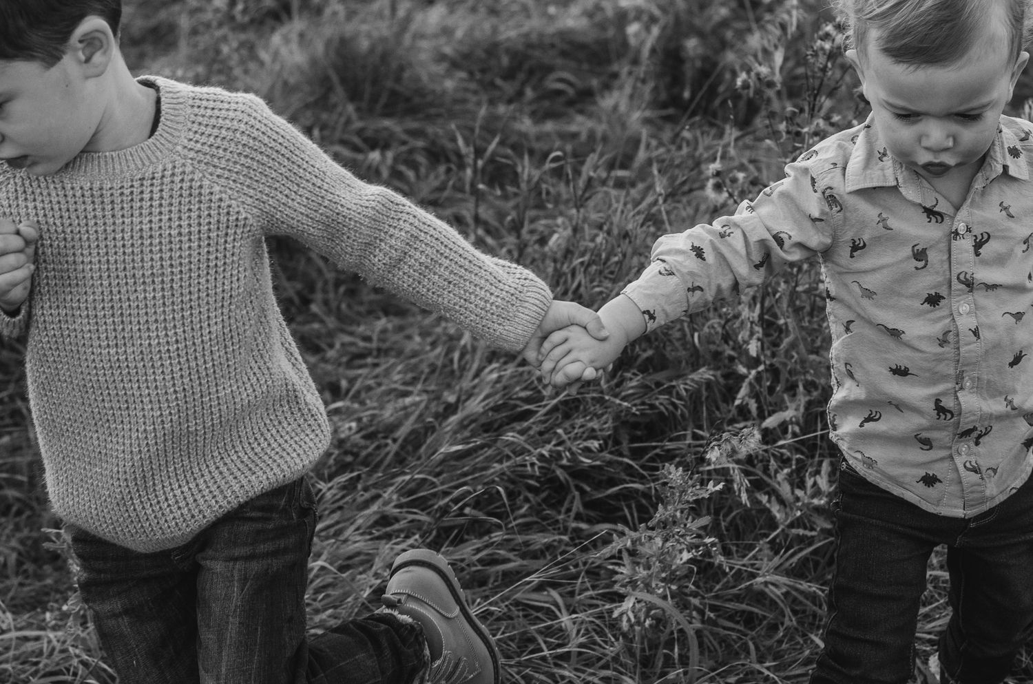 Two young boys hold hands walking through tall grass in a black and white outdoor photo.
