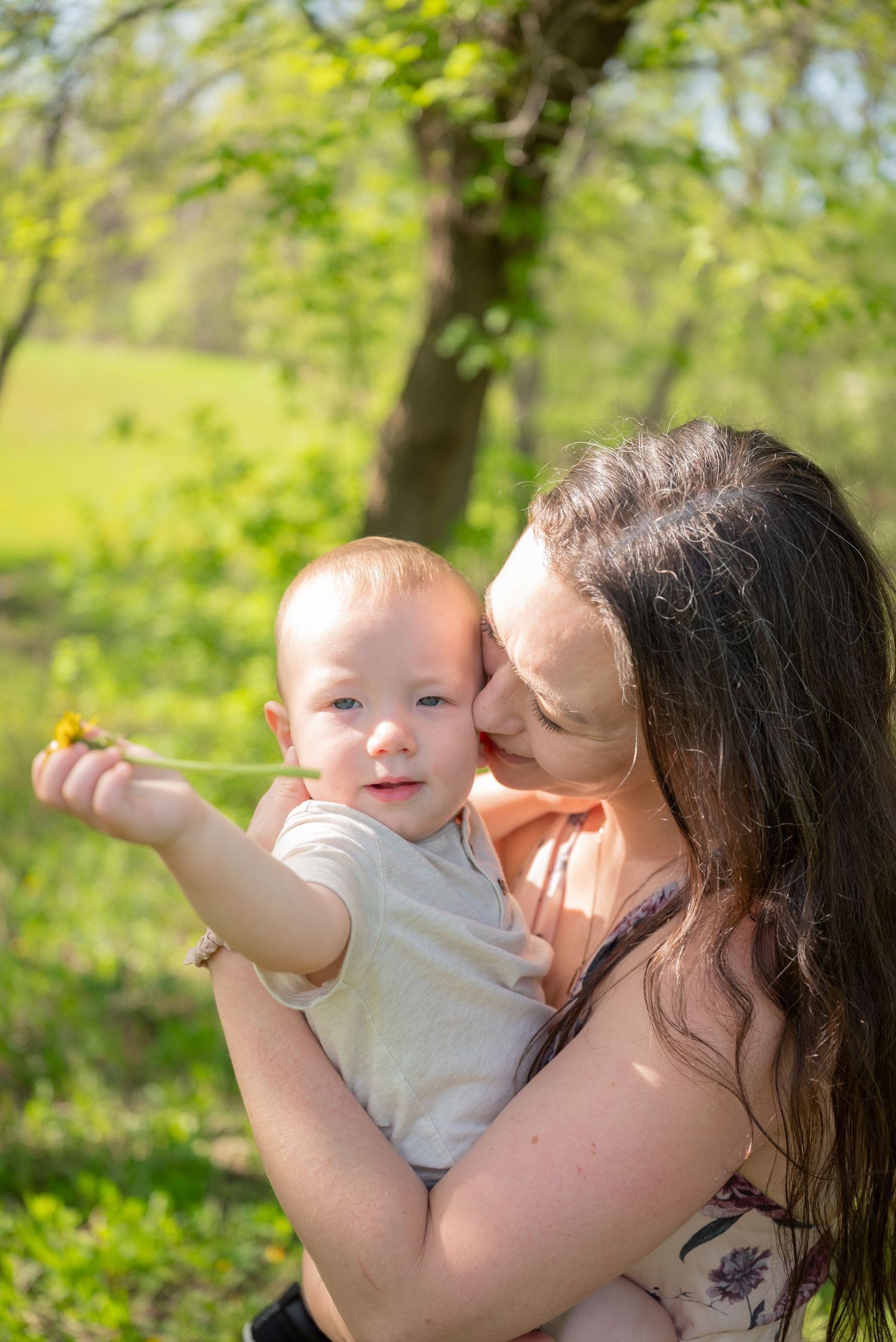 A loving mother kisses her baby boy outdoors while he reaches out holding a small flower in a sunny green park.