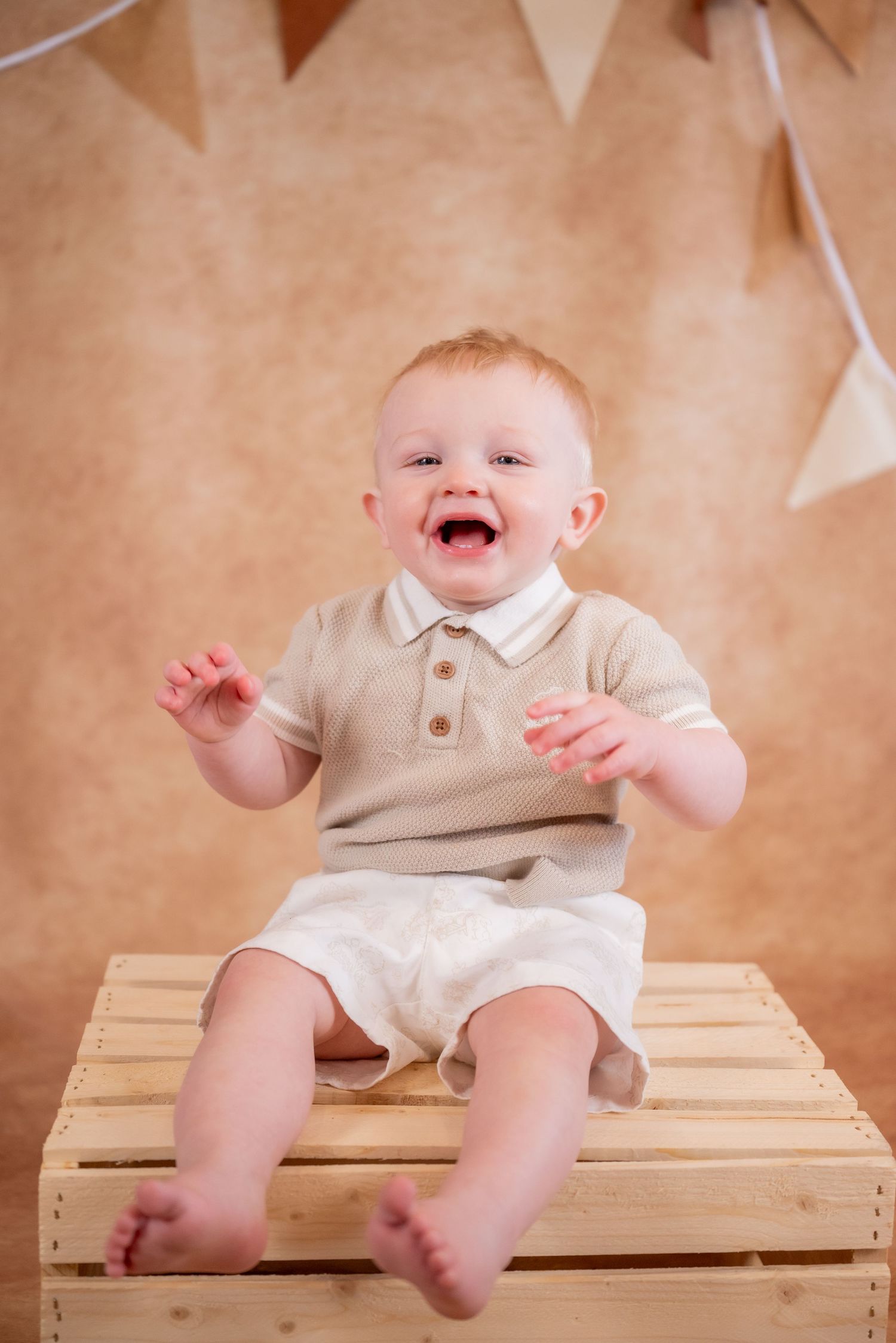 Happy laughing baby boy sitting on a wooden crate wearing a beige polo shirt and white shorts.