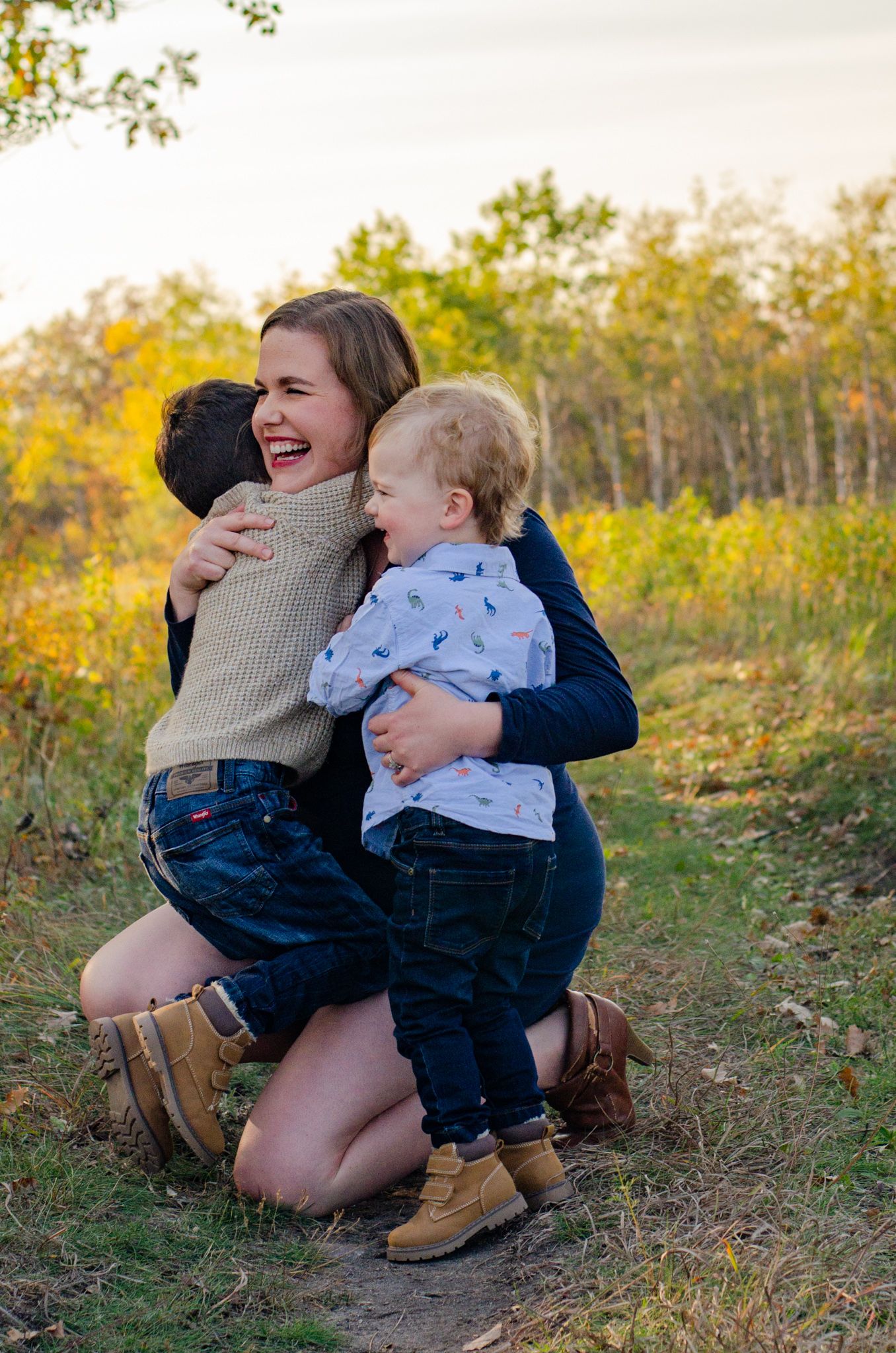 A laughing woman kneels outdoors hugging two young children in a golden autumn field.