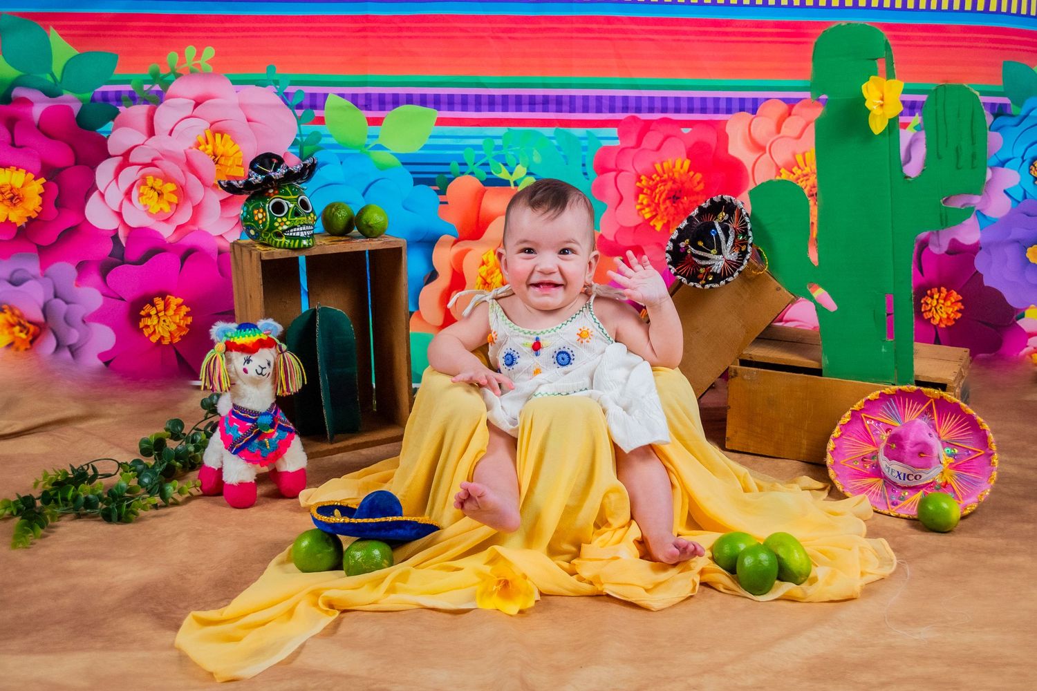 Baby smiling in colorful fiesta-themed photo shoot with paper flowers, cactus, maracas, and lime props on sandy background.