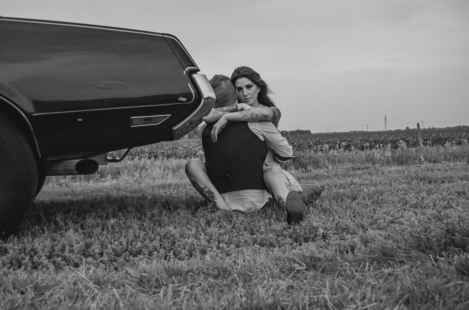 Black and white photo of a woman sitting in grass leaning against a vintage car in an open field.