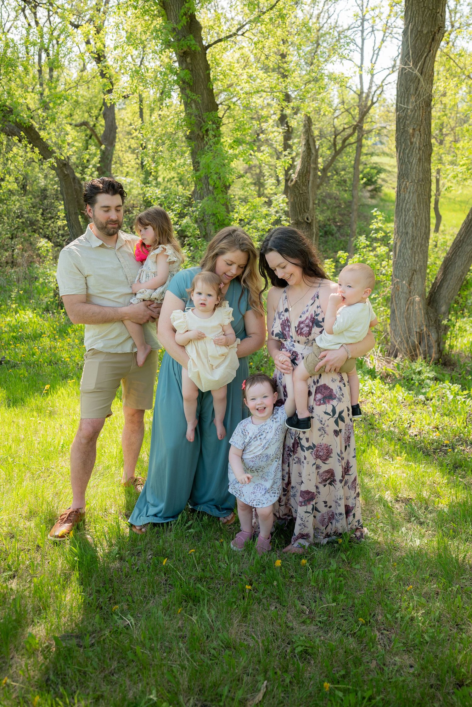 Family of seven poses outdoors in spring greenery, two mothers in floral dresses holding babies with toddlers nearby.