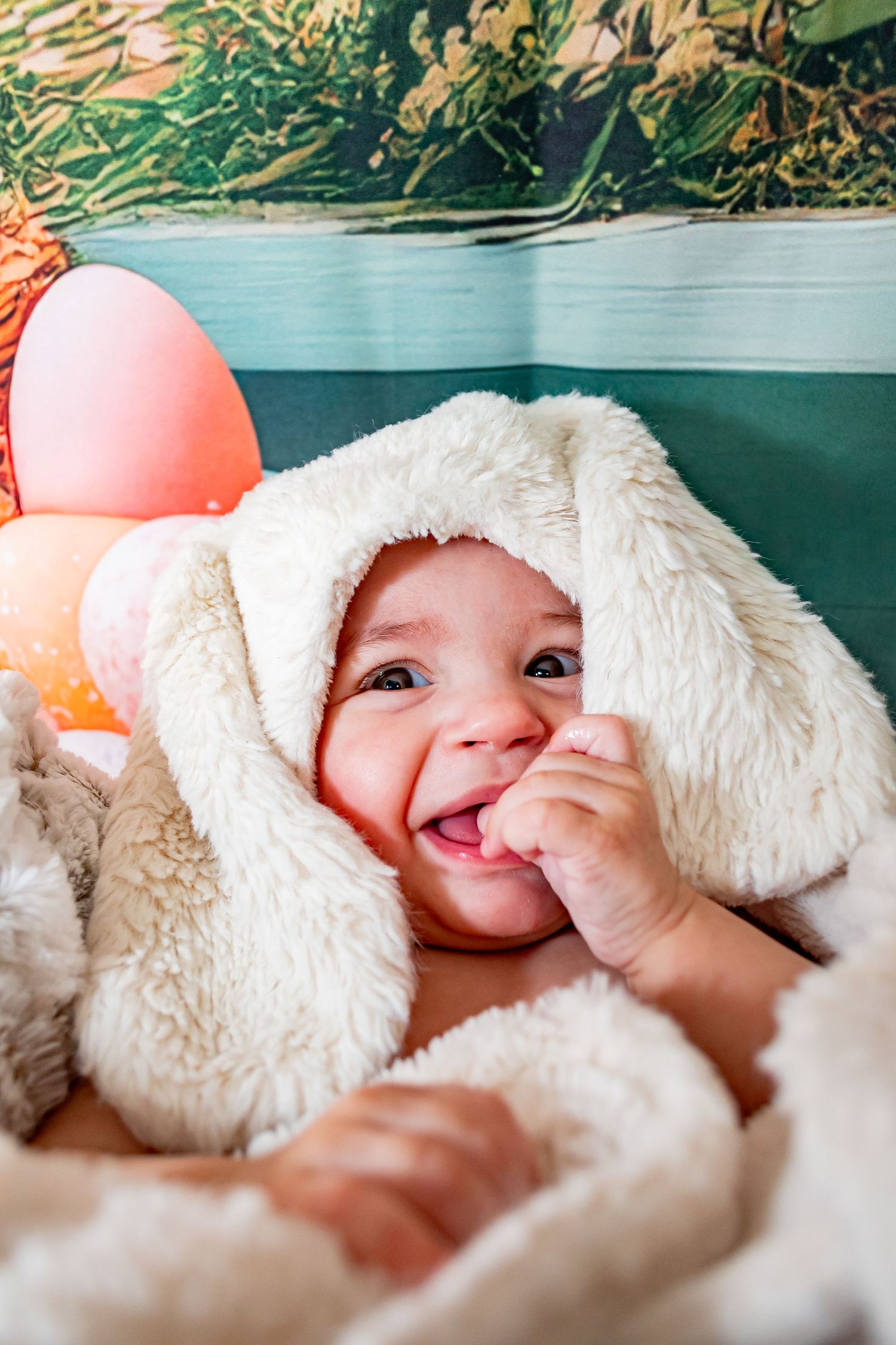 Smiling baby in a white fluffy bunny costume with floppy ears, surrounded by colorful Easter eggs.