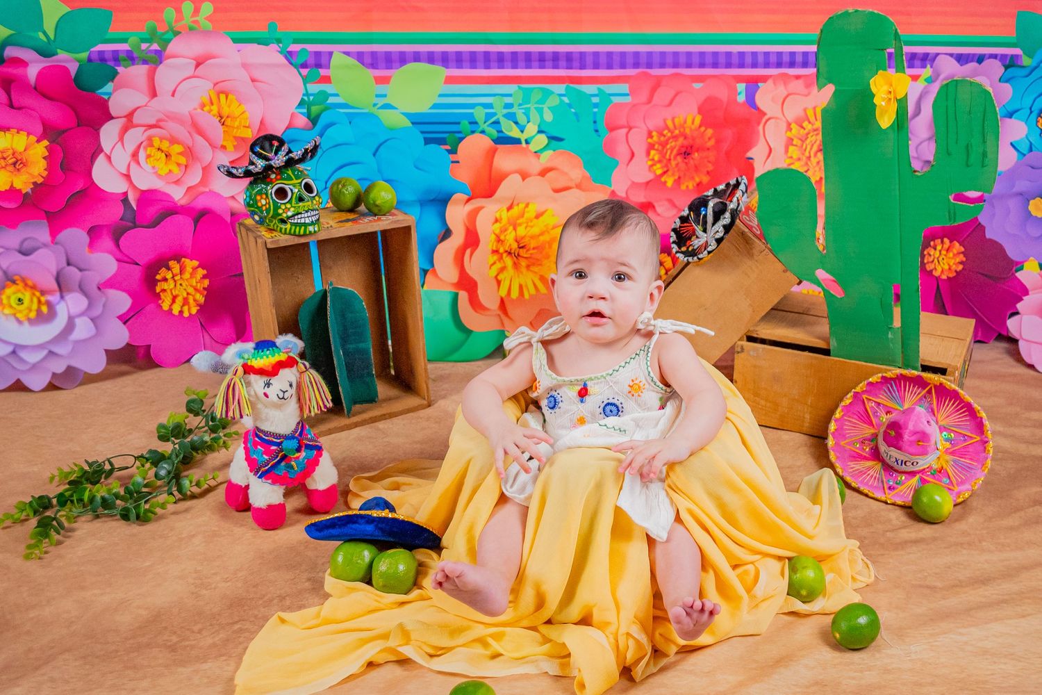 Baby girl in yellow dress sits on sand surrounded by sombreros, limes, and colorful floral fiesta backdrop decorations.