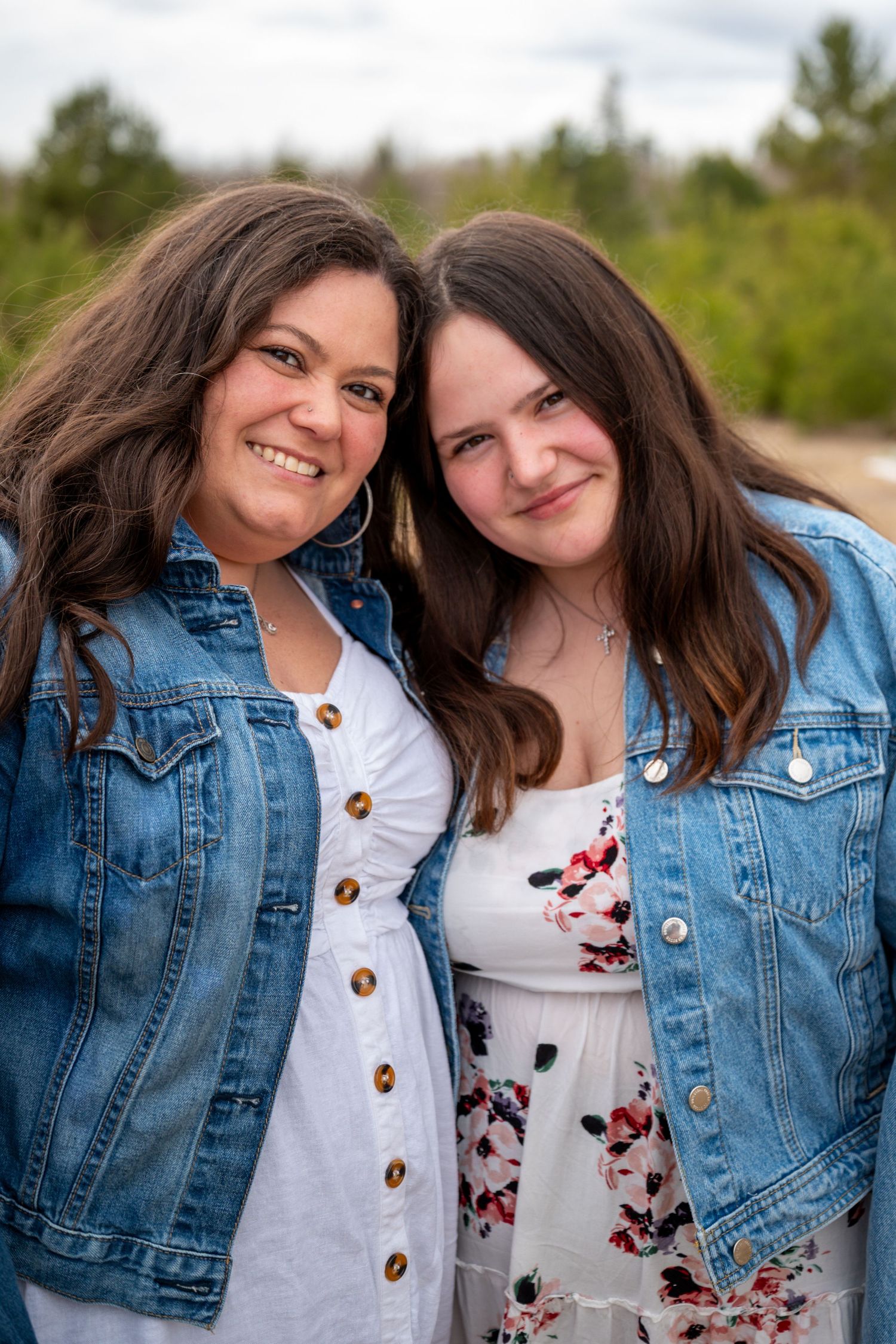 Two women in white dresses and denim jackets smile together outdoors with trees in the background.