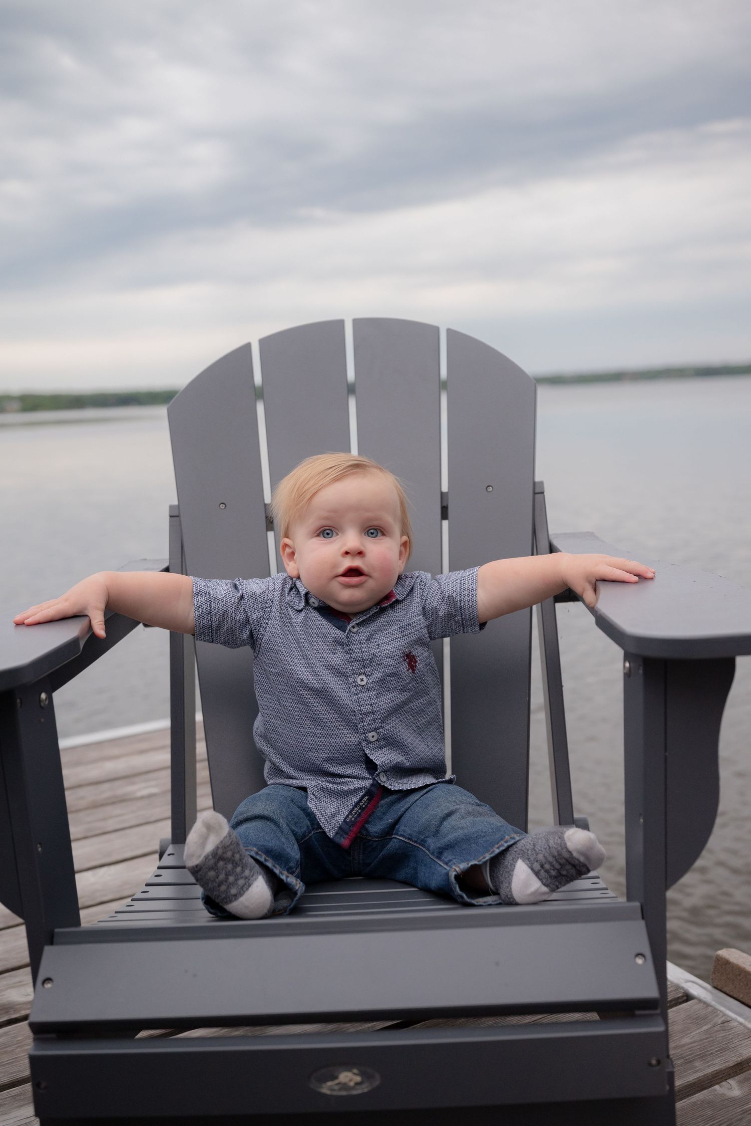 Cute toddler boy sitting in a gray Adirondack chair on a wooden dock with arms outstretched over a lake.