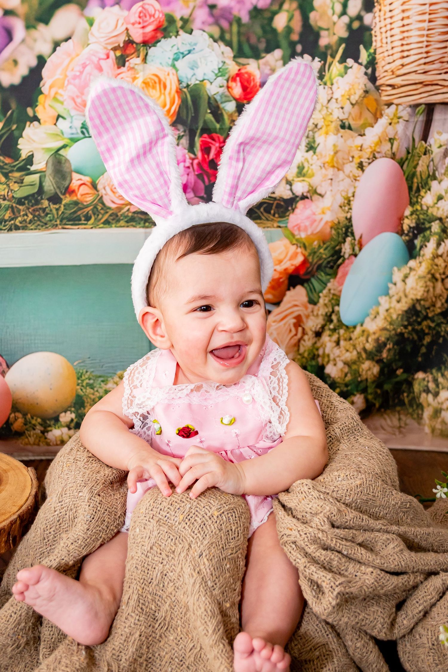 Smiling baby wearing pink bunny ears headband and pink outfit, sitting on burlap with Easter eggs and floral backdrop.
