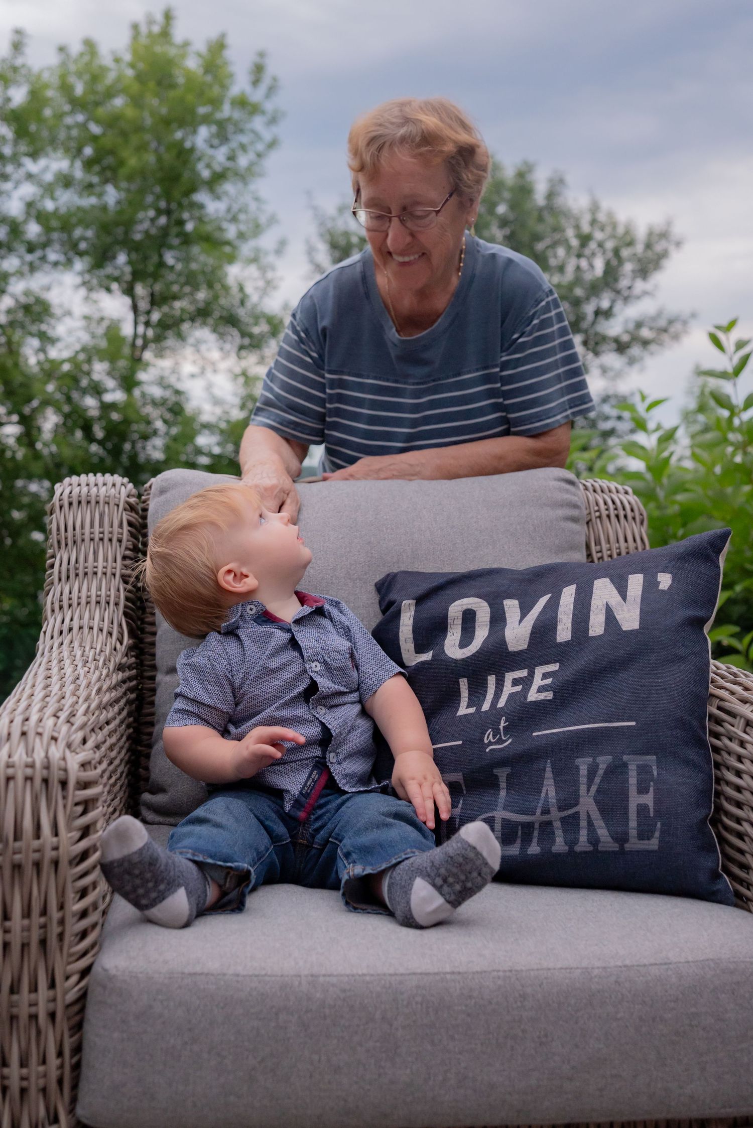 Elderly grandmother smiling at toddler boy sitting together on wicker chair outdoors with Lovin Life at the Lake pillow.