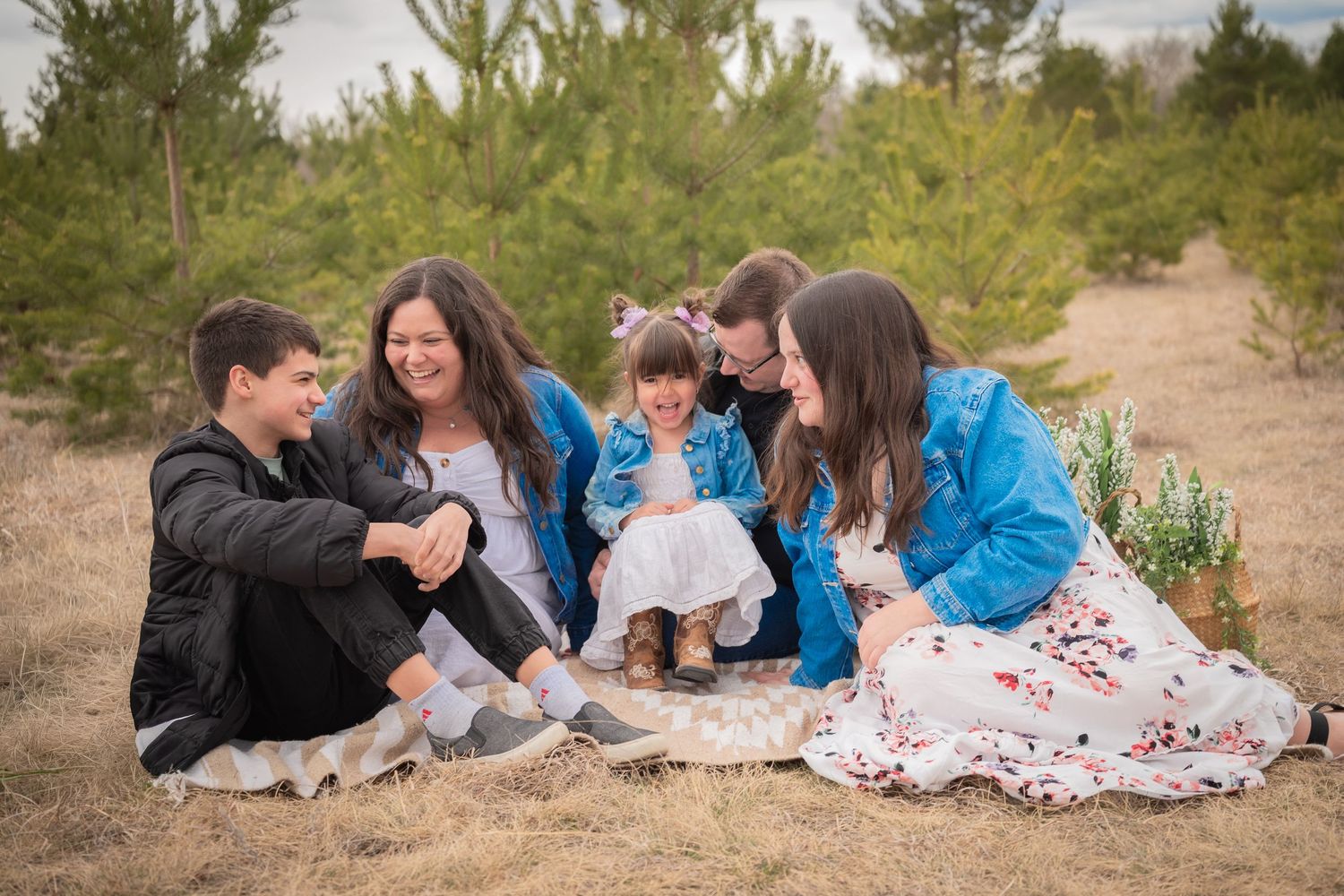 Happy family of five sitting together outdoors in a pine forest setting, laughing and bonding on dry grass.