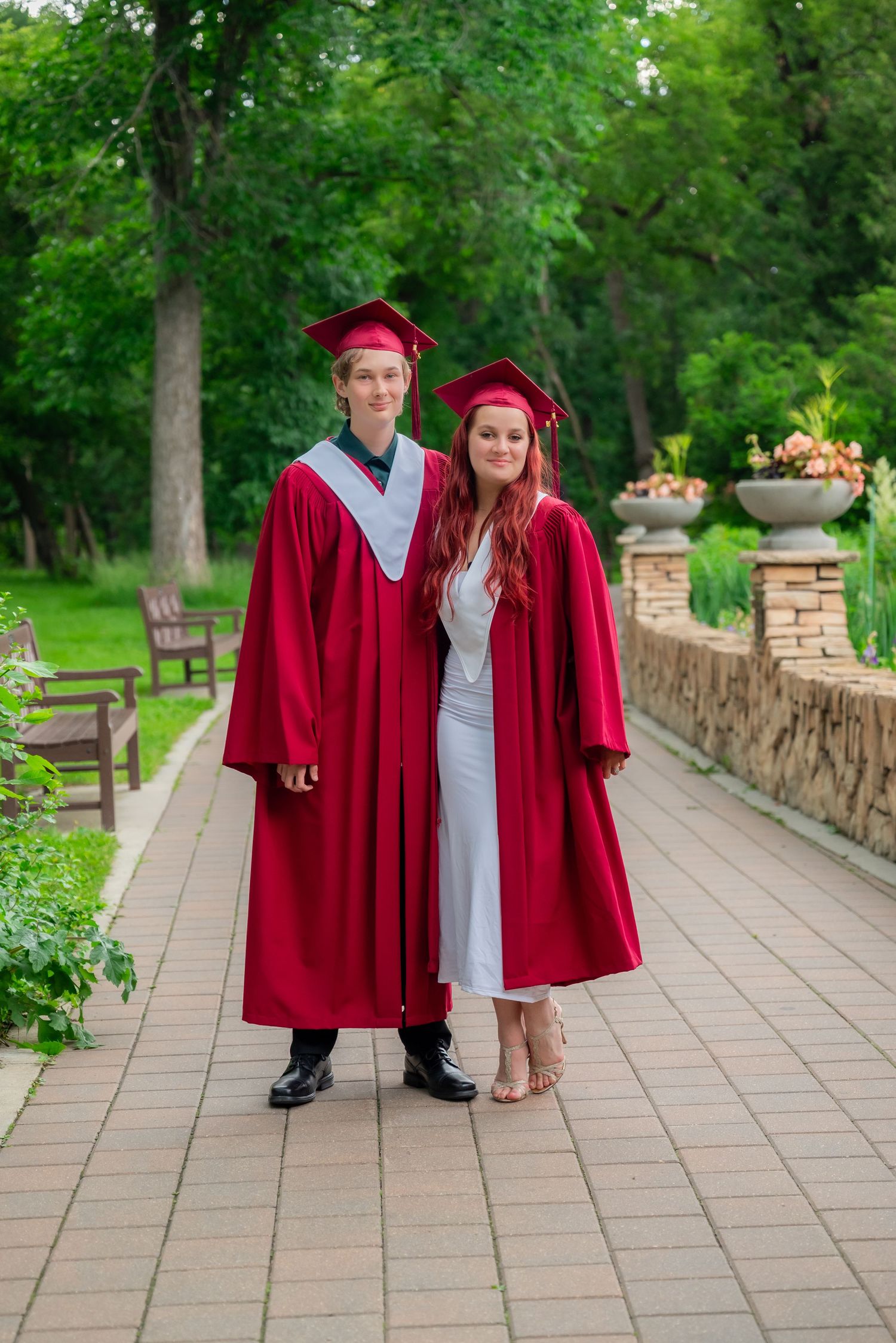 Two graduates in red caps and gowns pose together on a garden pathway surrounded by lush green trees.