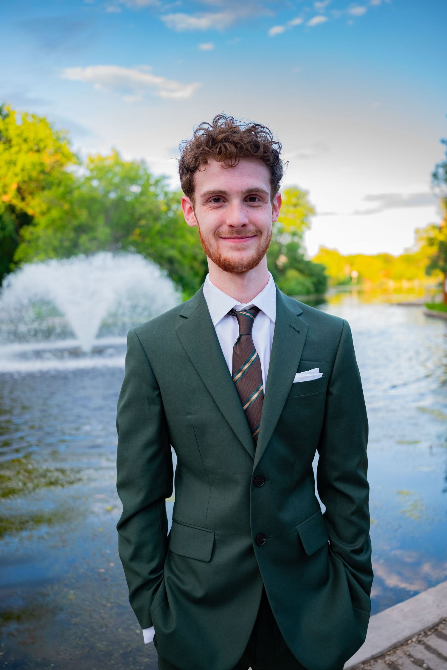 Young man in green suit and striped tie smiling outdoors near fountain and trees.