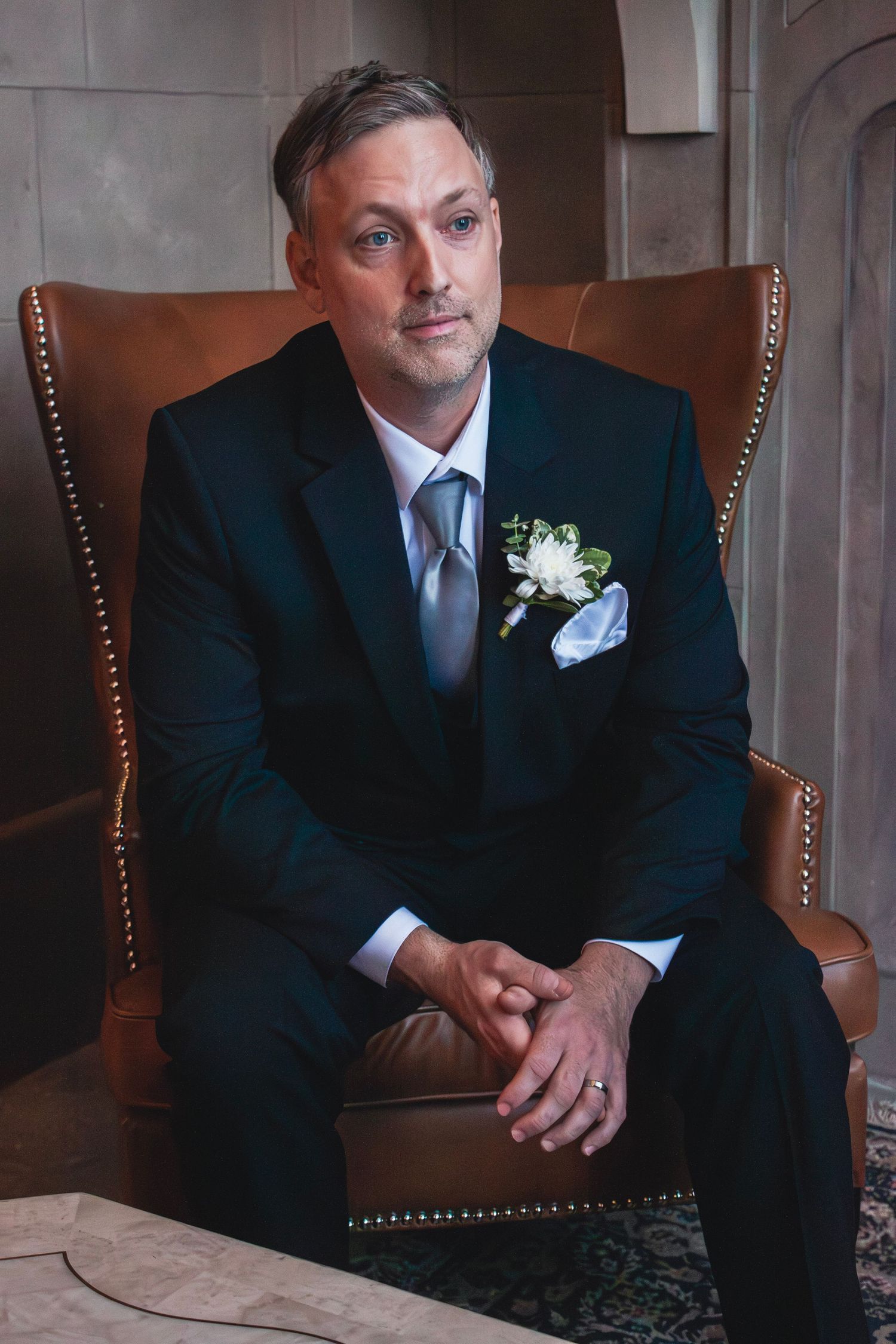 A groom in a dark suit with white boutonniere and gray tie sits in a leather wingback chair, hands clasped.