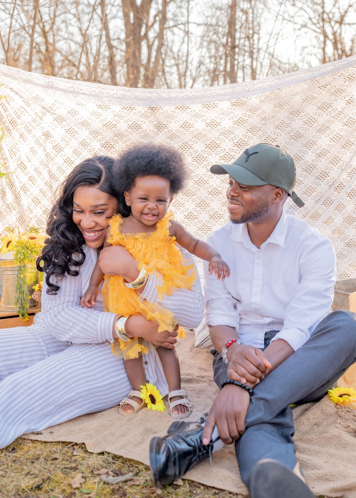 A joyful Black family with a toddler in a yellow dress sits on a blanket outdoors with a lace backdrop.