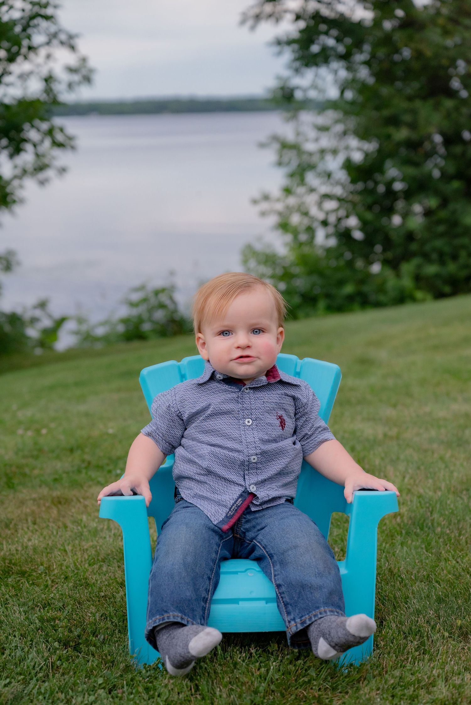 Toddler boy in blue jeans and gray shirt sitting in a turquoise Adirondack chair on green grass by a lake.