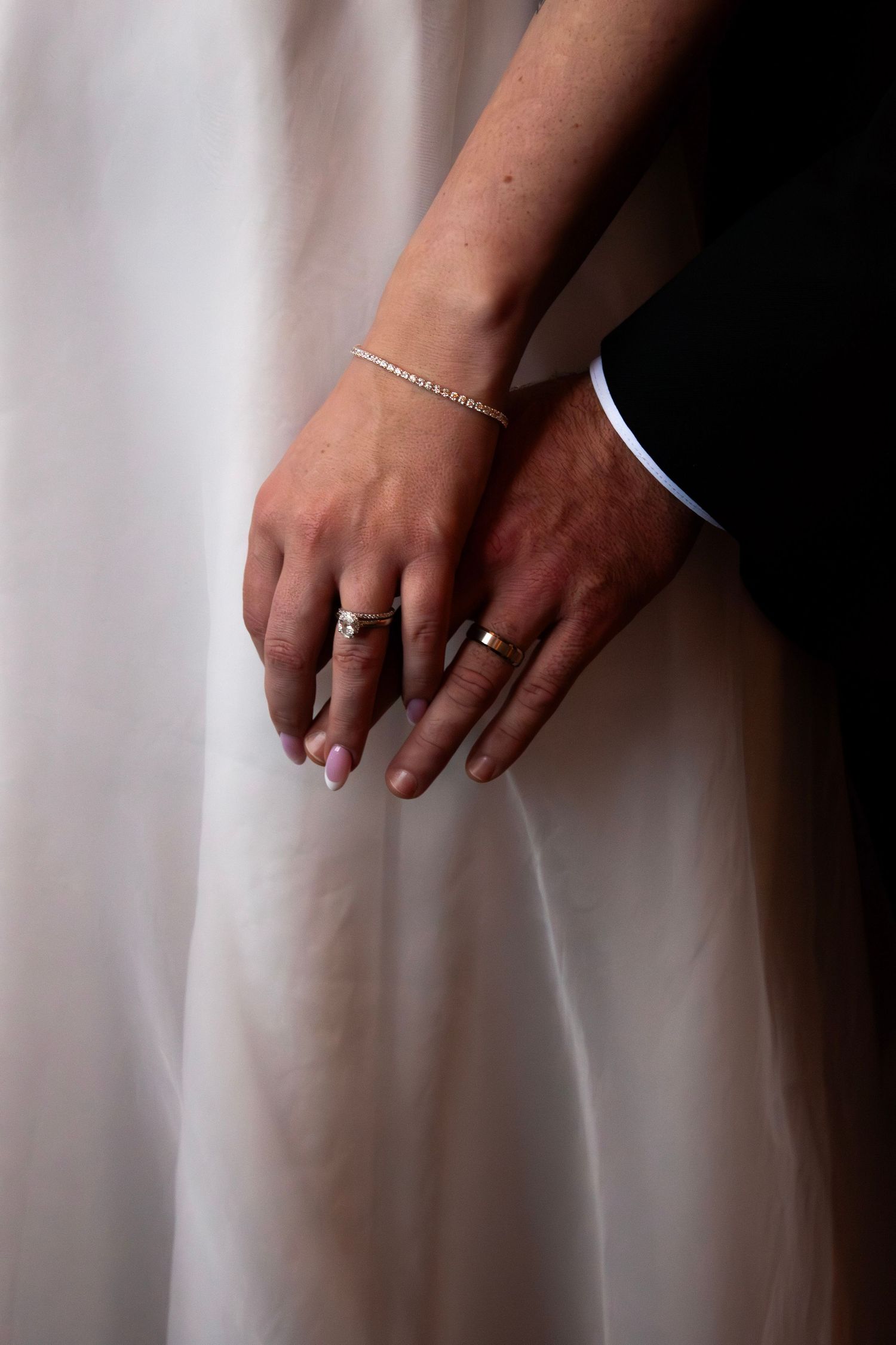 Newlyweds hold hands showing wedding rings, bride wearing a diamond bracelet and white gown.