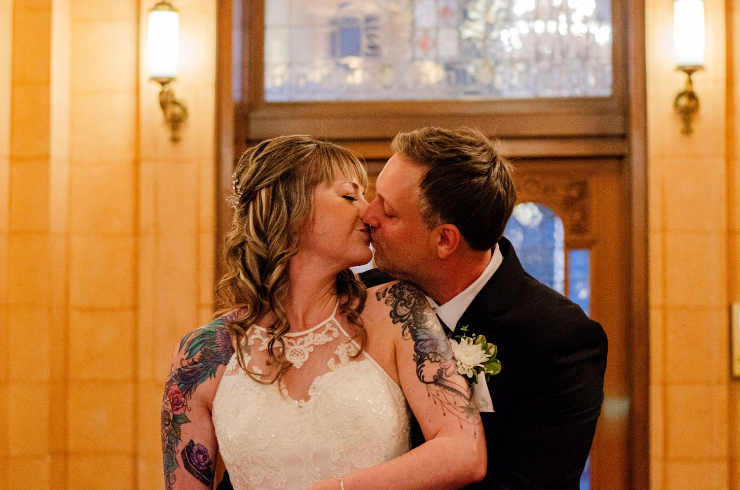 Bride and groom share a romantic kiss at their wedding venue, bride wearing a white lace gown with tattoos visible.