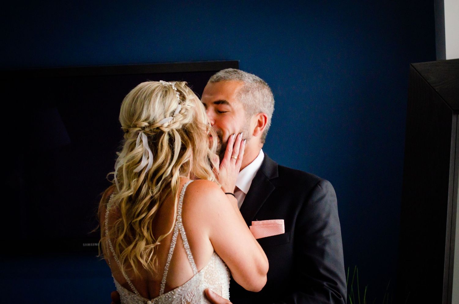 Bride and groom sharing a romantic kiss, with the bride in a lace gown against a navy blue wall.
