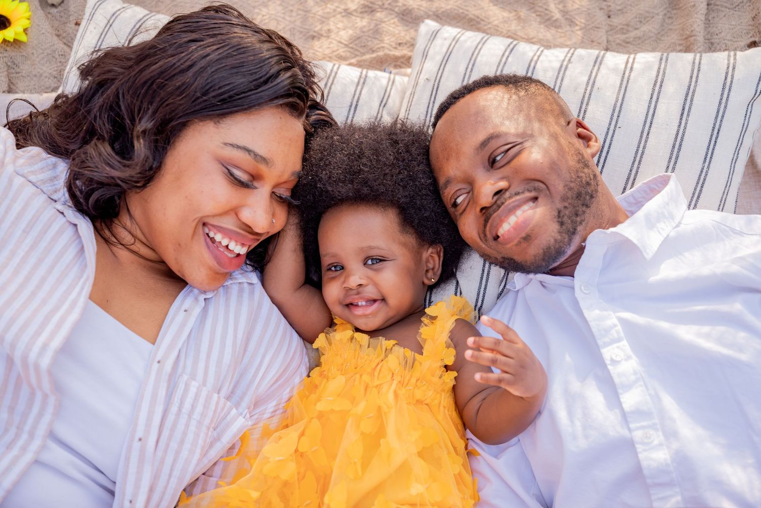 A joyful Black family of three lying together, parents smiling at their toddler daughter in a yellow dress.