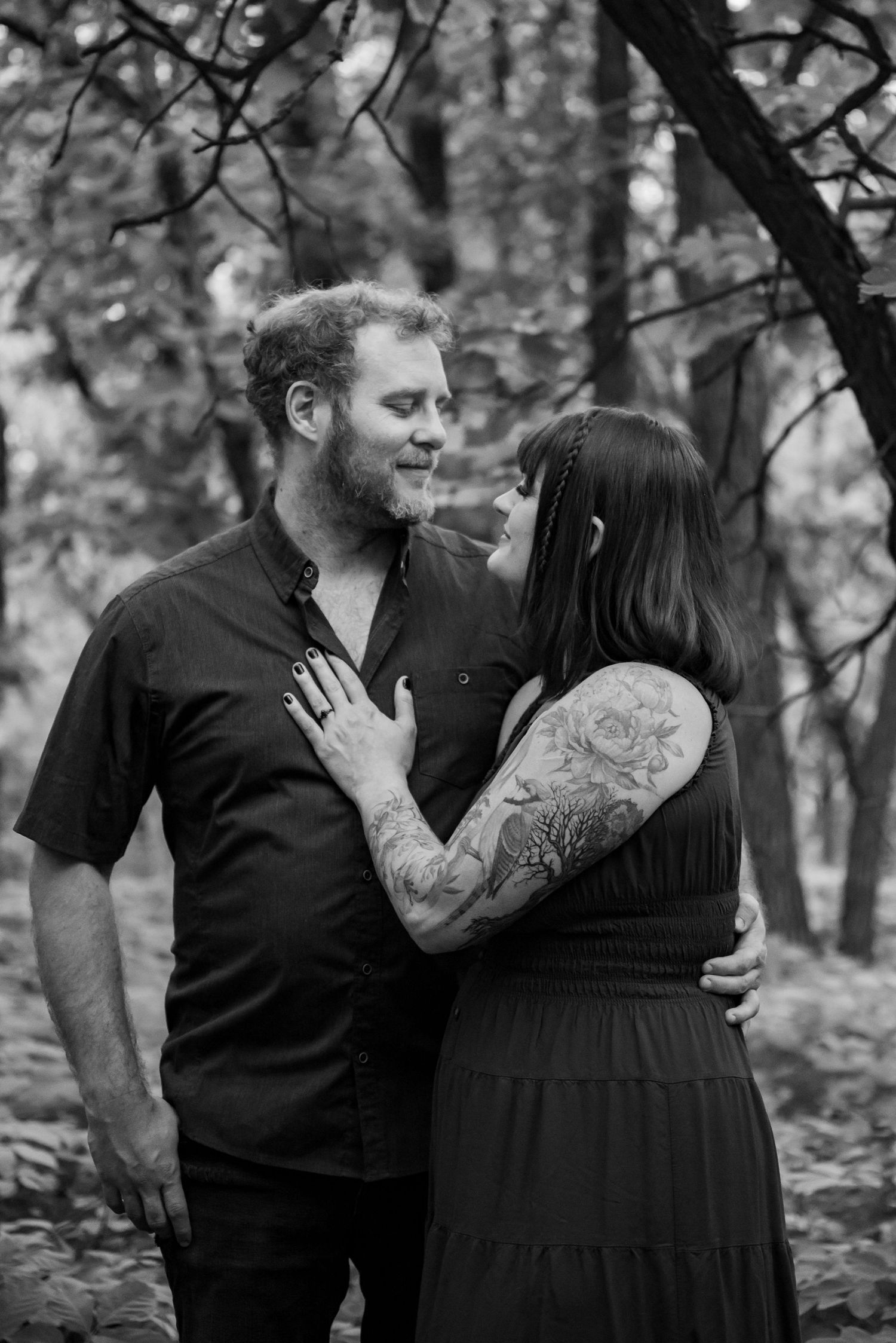 Black and white photo of a tattooed couple sharing a romantic moment in a forest setting.