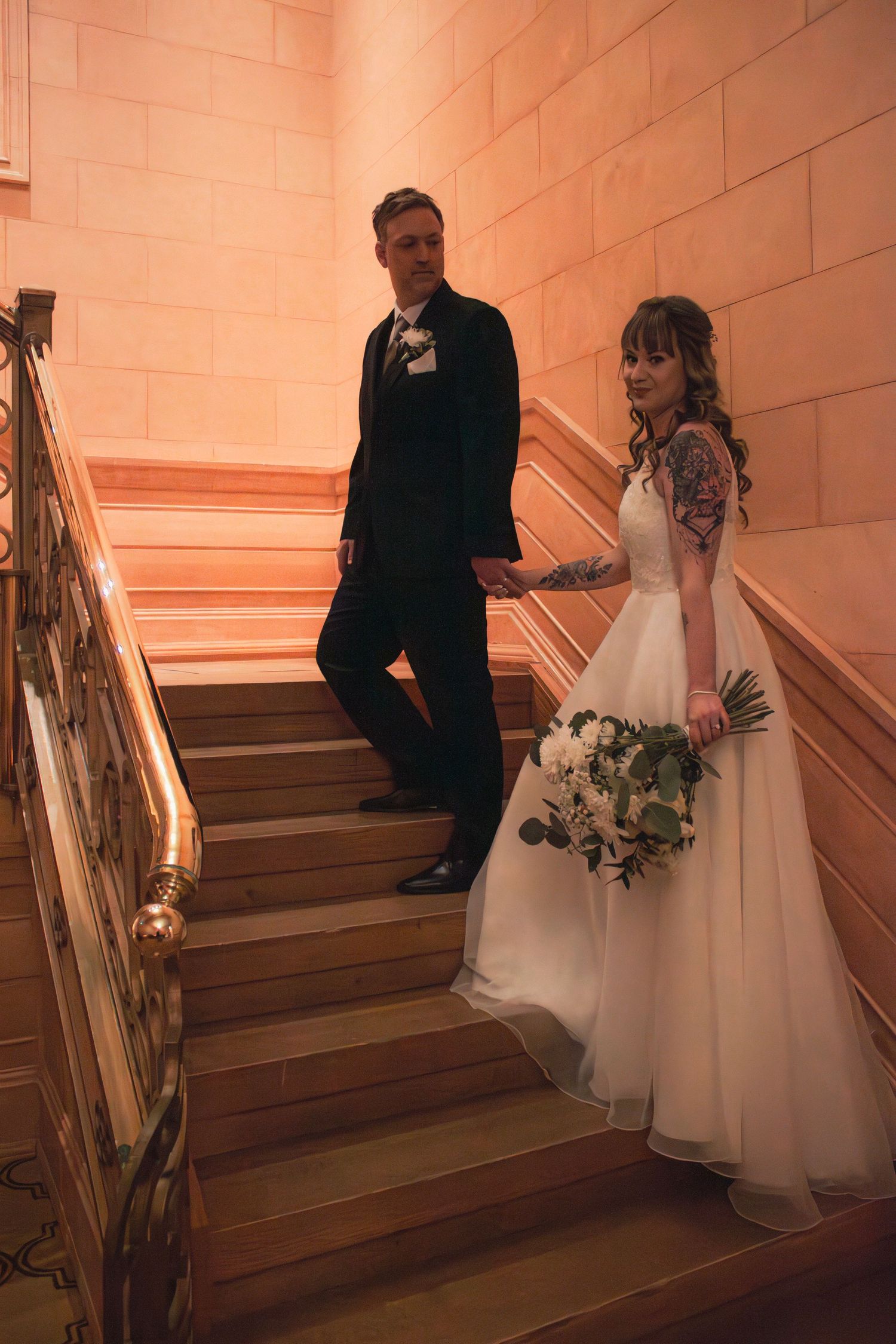 Bride and groom pose on elegant staircase, bride in white gown holding bouquet, groom in black tuxedo.