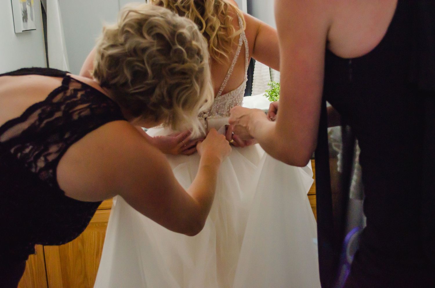 Two women helping a bride button up her white wedding dress from behind in a warmly lit room.