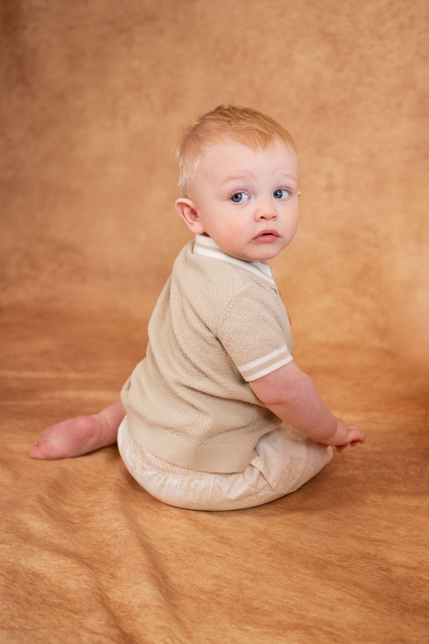 Adorable baby with blue eyes sitting on a tan background wearing a beige outfit, looking over shoulder.
