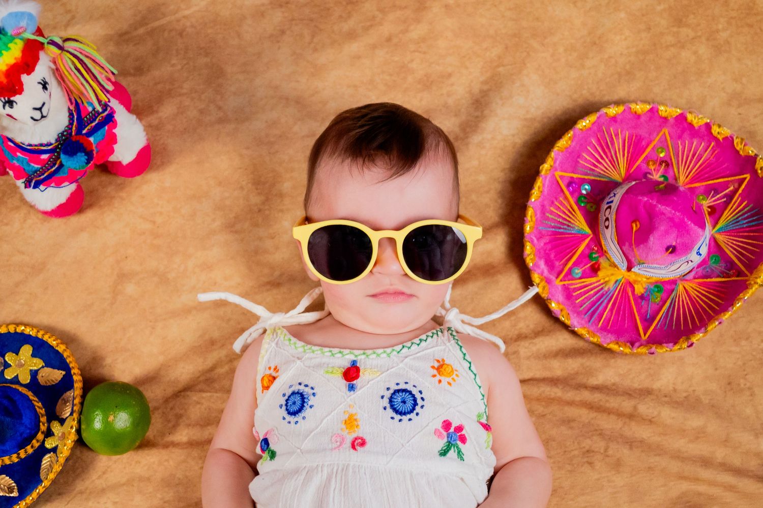 Cute baby wearing yellow sunglasses lying next to colorful mini sombreros and toys on a tan background.